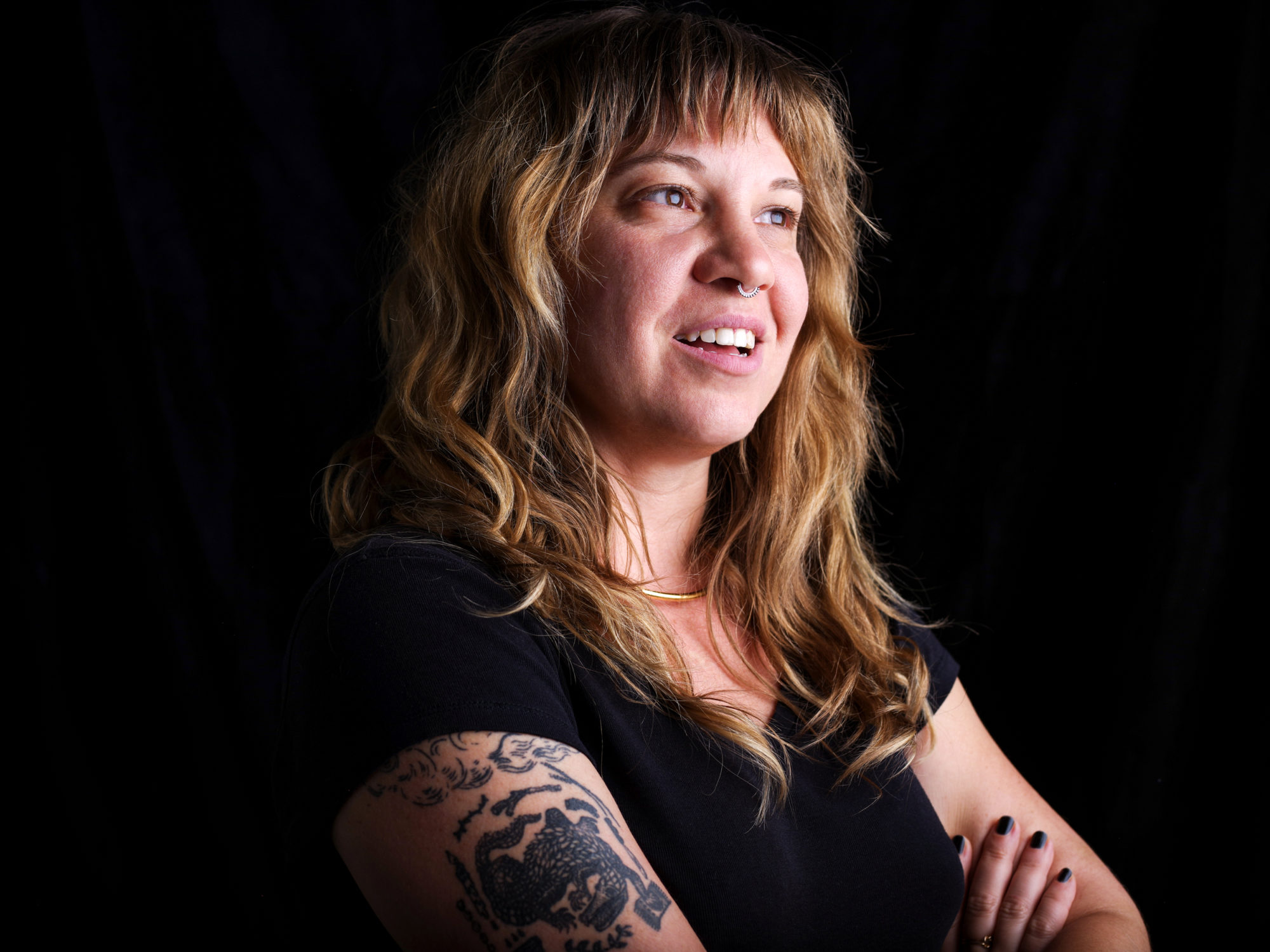 woman with wavy hair and tattoos crossing her arms in a black shirt against black background.