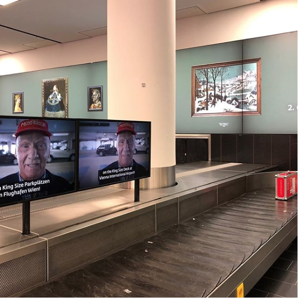 two screens with the same image of an old man in a red hat above a baggage belt.