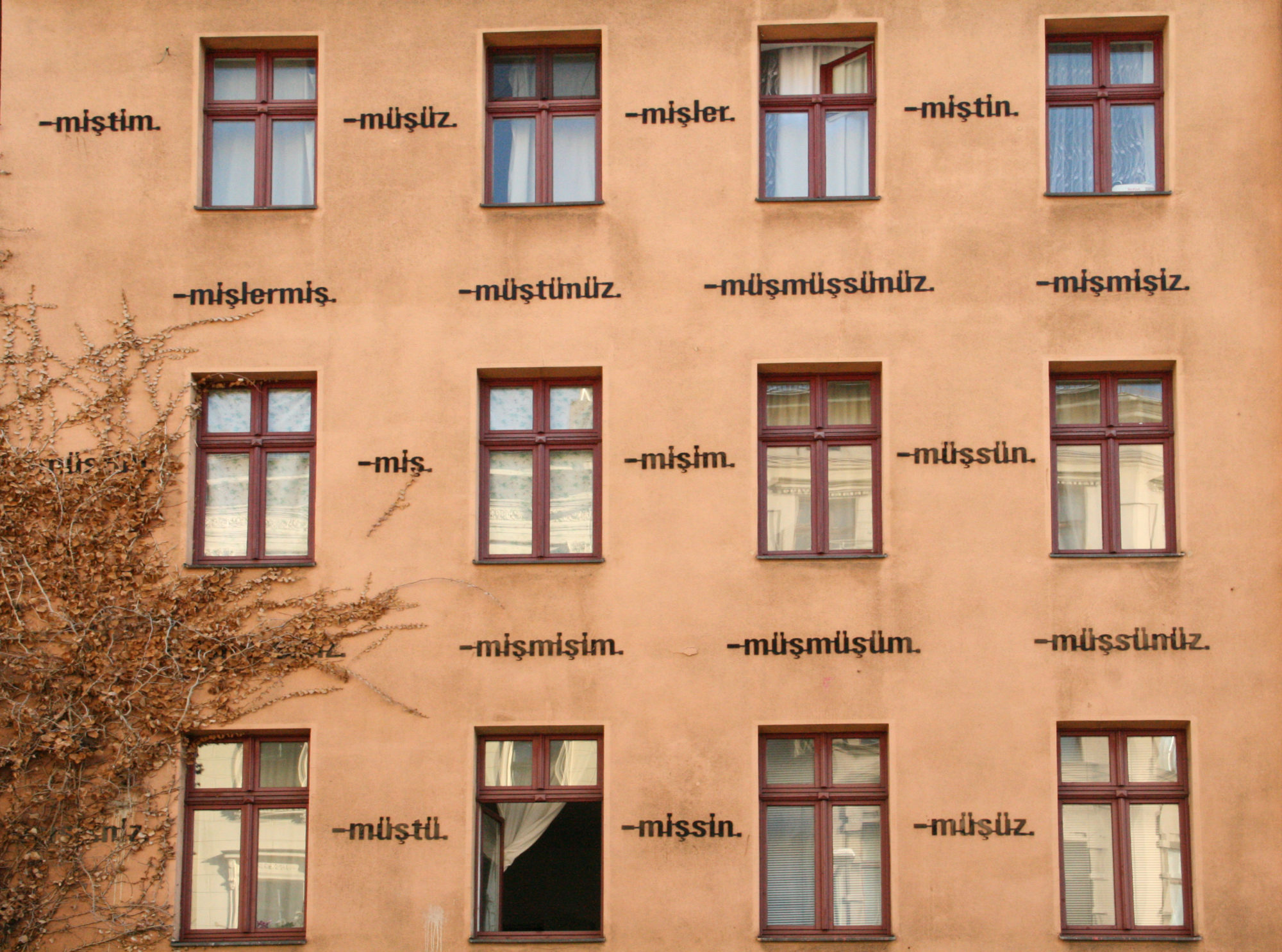 a tan building with red-framed windows. an orange vine grows along the side of the building, and several of the windows have blown open