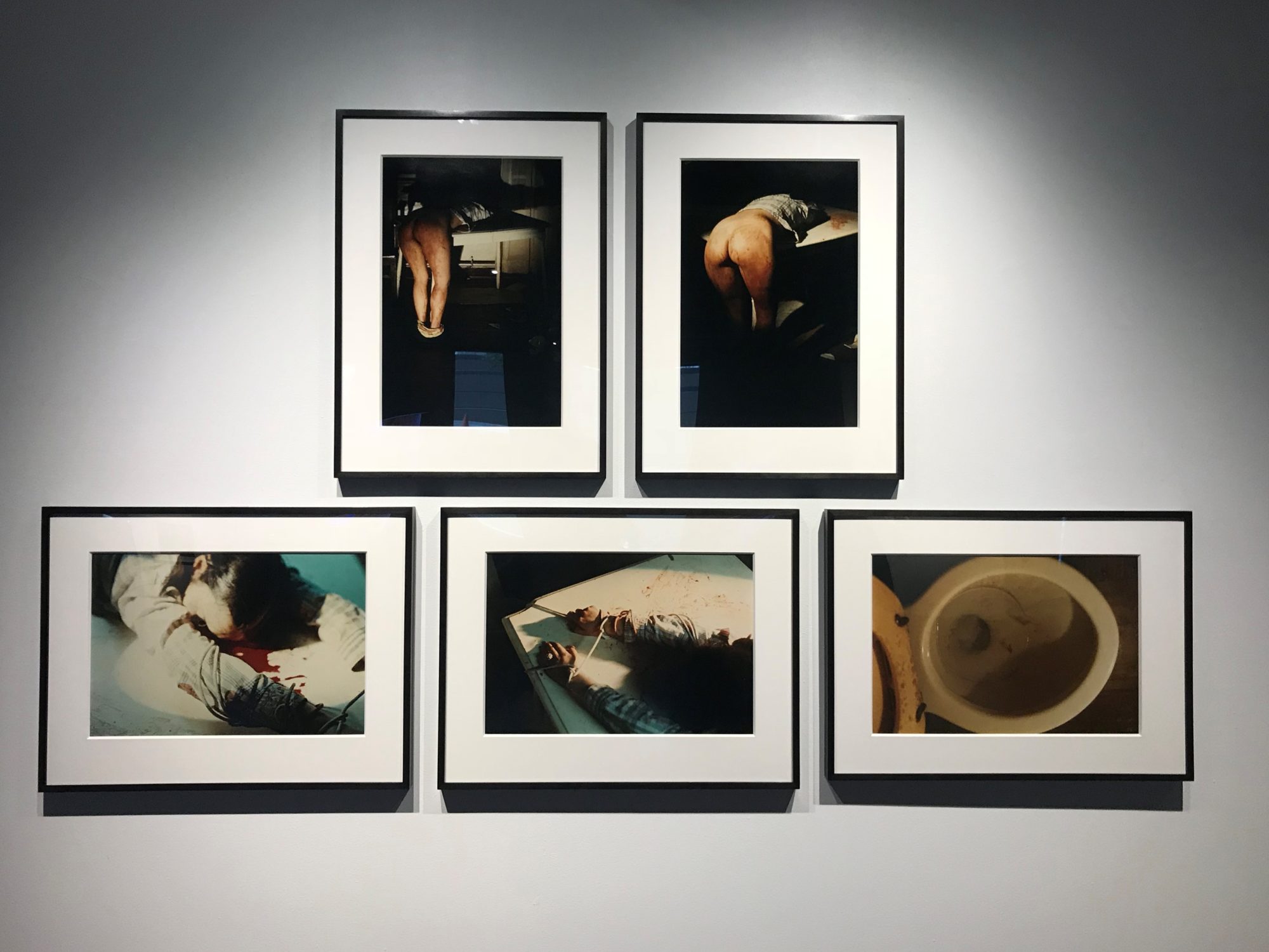 *Content Warning* Five photographs are hung on a white wall: four depict a woman, smeared in blood and naked from the waste down, tied to a wall; one depicts the inside of a toilet bowl.