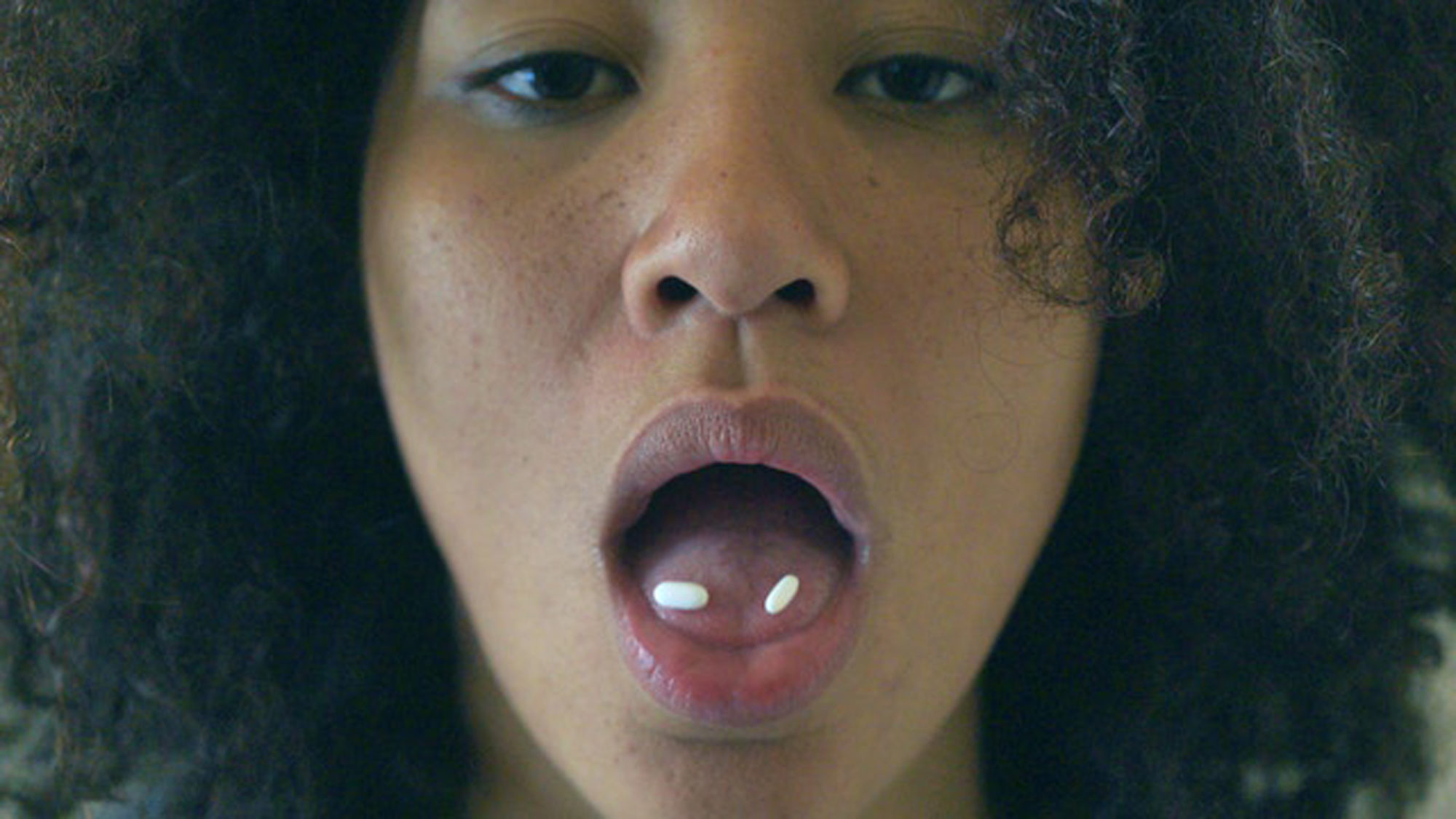 A close-up photograph of a woman looking straight into the camera, her mouth open and two white pills resting on her tongue.