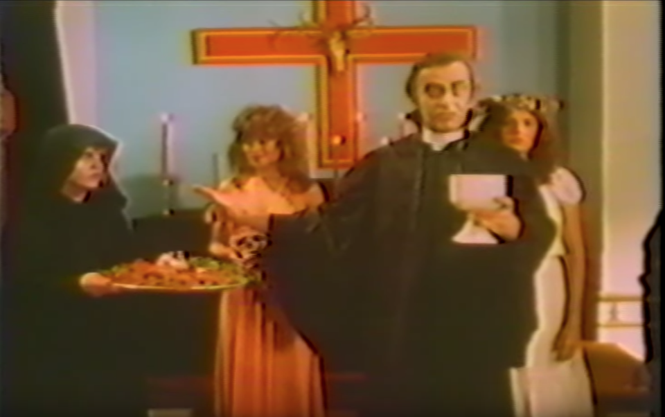 A priest in a church, standing before an altar, holds a chalice. Three women stands behind him: a woman in a black robe holding a tray of food, a woman in an orange dress holding a skull, and a woman in a white dress looking listlessly forward.