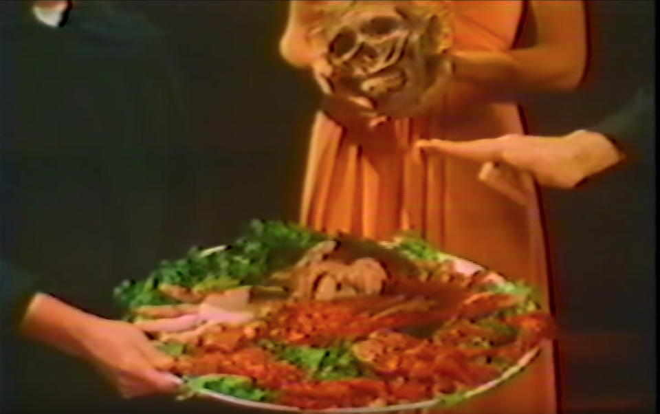 A woman in the center, wearing an orange dress, holds a human skull. A figure to the left holds a circular tray of food. A hand, emerging from the right side of the frame, hovers over the dish of food.