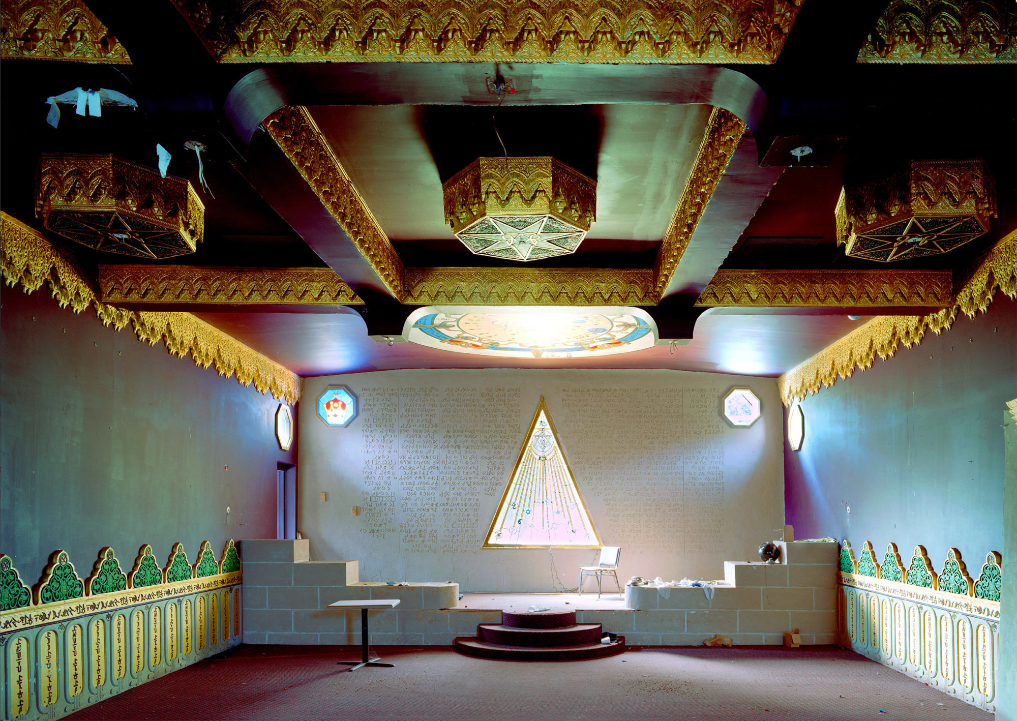 an ornate room with a colorful glass ceiling window, a stage, and a glass triangle window that is central. the room is lined with interesting baseboard patterns, the ceiling is lined with intricate gold design