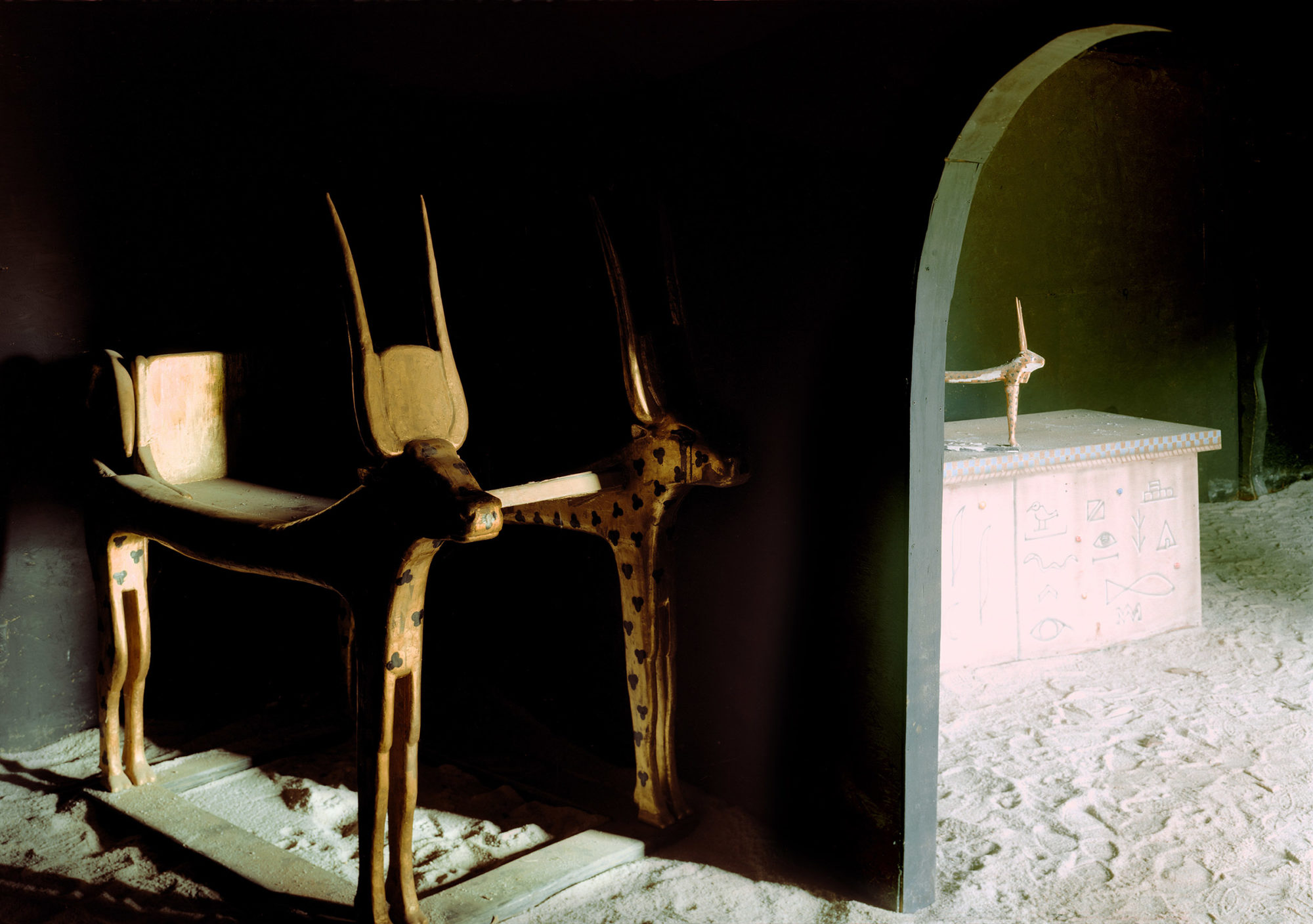 dimly lit green interior space with sand floor and Egyptian-looking wooden animal sculpture