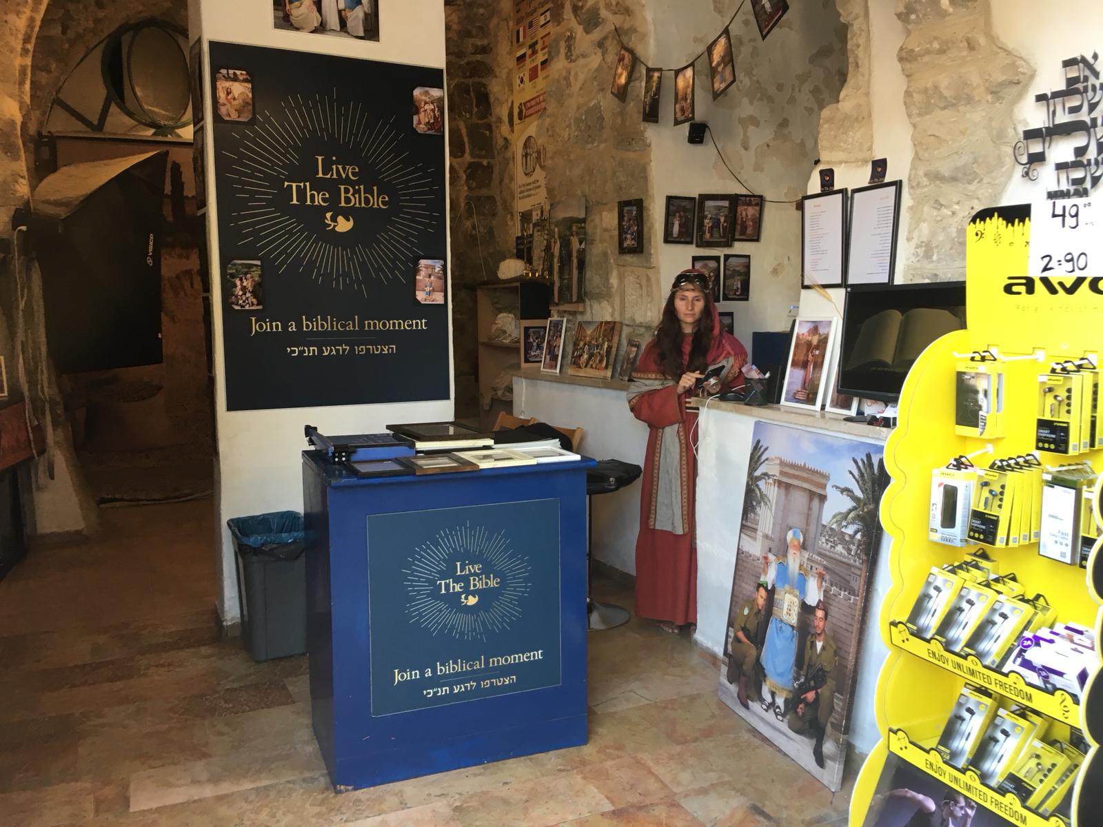 A woman stands in a gift shop selling headphones and phone cases, along with biblical images. A poster on a wall advertises, 