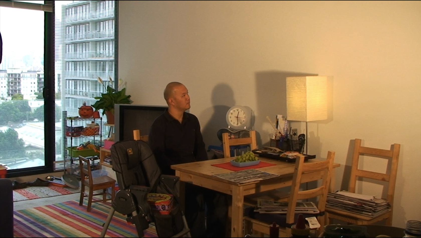 a man sitting at a wooden table with grapes in the center, and chairs littered with magazines. there is a booster seat for a child who is not present, and the man has a far-away look in his eyes, as though lost in thought