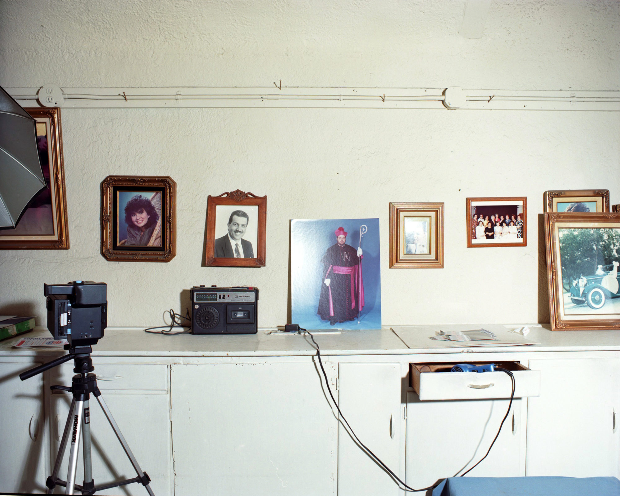 A series of portraits hang against white wall with camera equipment in the foreground