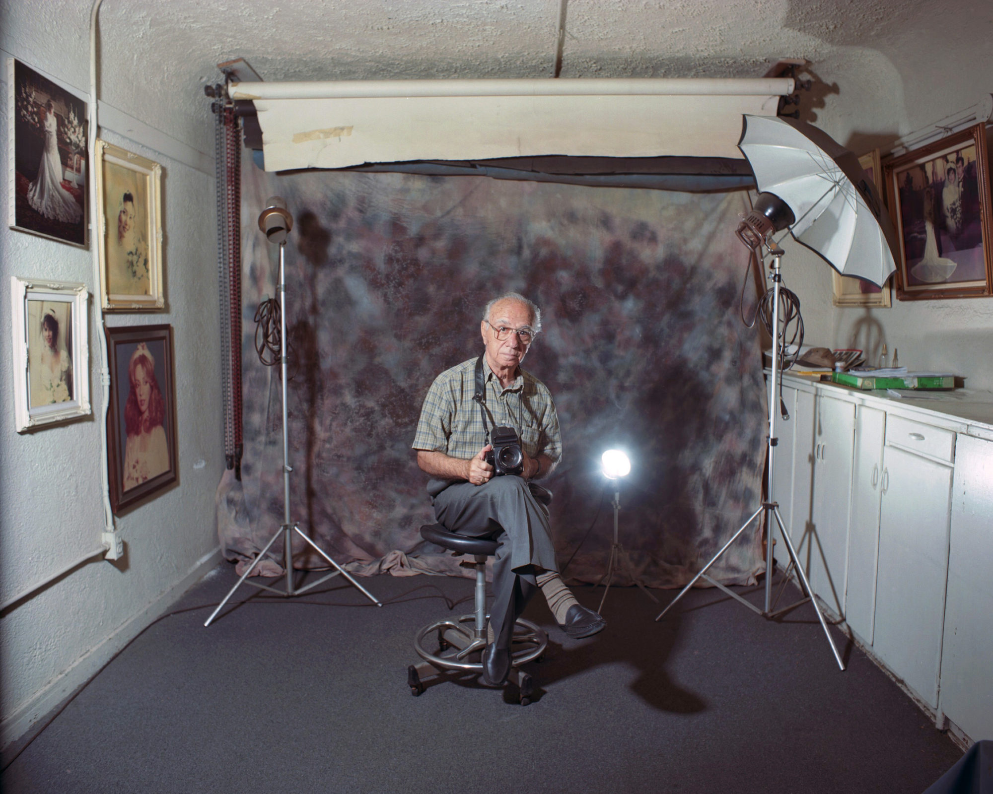 A portrait of an elderly man holding a camera in front of a tie dye backdrop in a room with hanging wall portraits