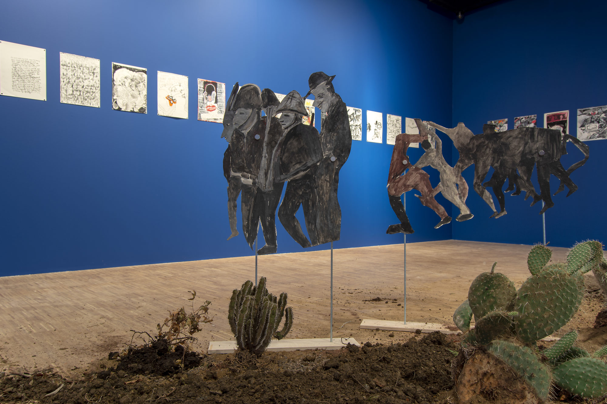 cut outs of cartoon men and beasts positioned over dirt and cacti in an installation