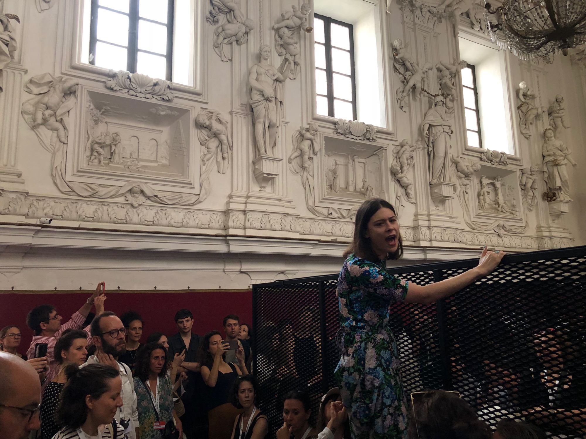 a woman in a floral print dress holding onto to the side of a black fence. a crowd bows their heads and listens, some watch her.