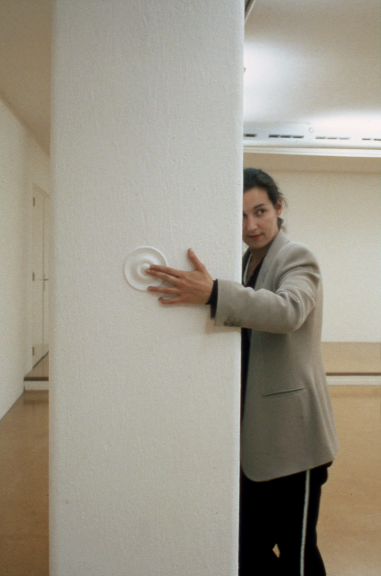 woman in taupe blazer leaning against white pillar and touching white circular pattern on the pillar