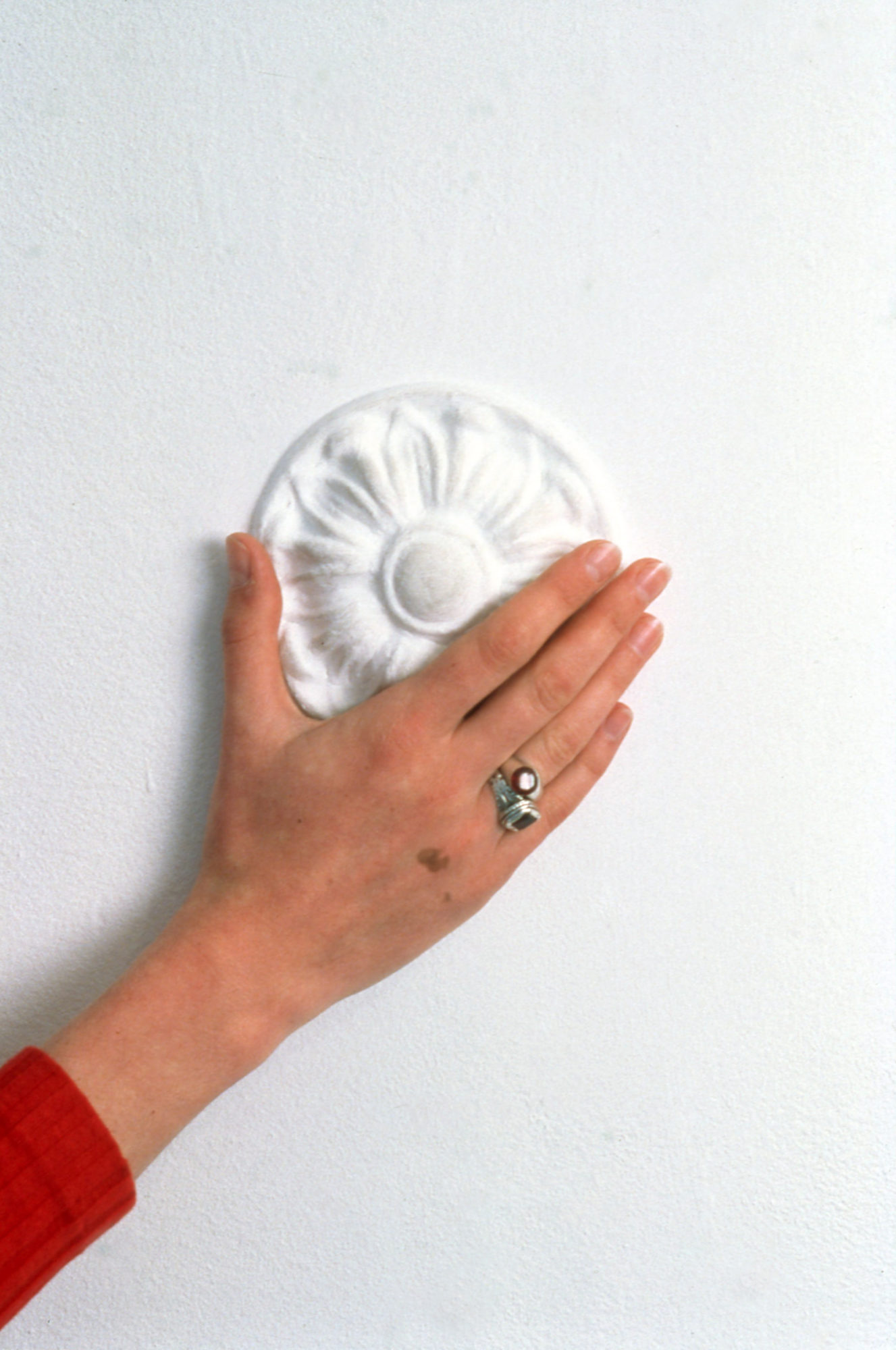 hand with two rings on the ring finger placed on top of a white ceramic circular pattern on a white wall