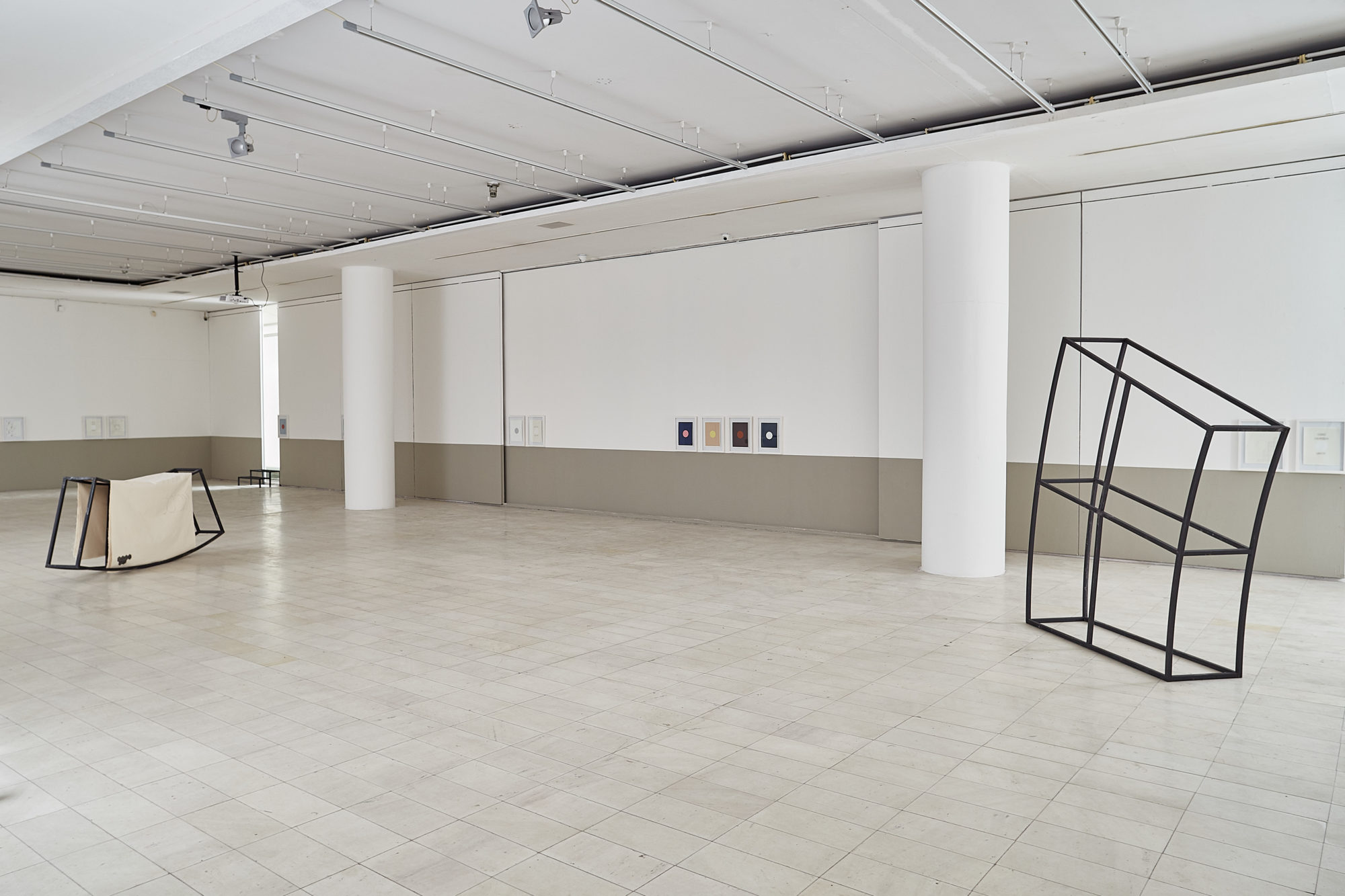 two black distorted rectangular sculptures, one with a cream fabric pulled over, in a white spacious room with small art pieces placed on the walls