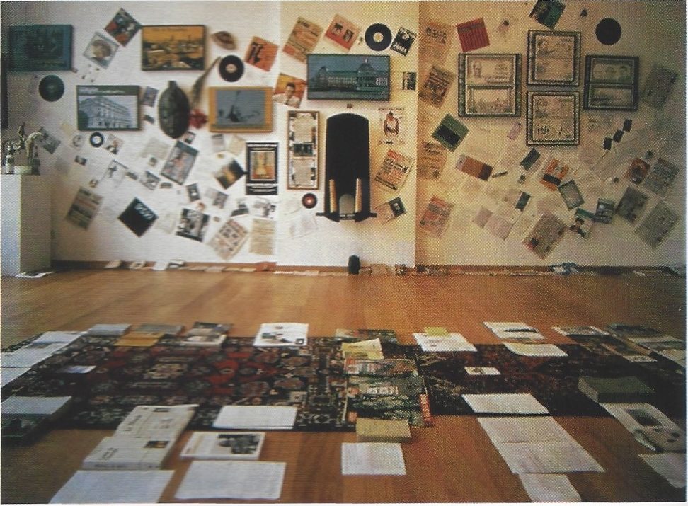 photo of a room filled with documents and a long rug on wood floor and a large arrangement of multiple objects on a white wall