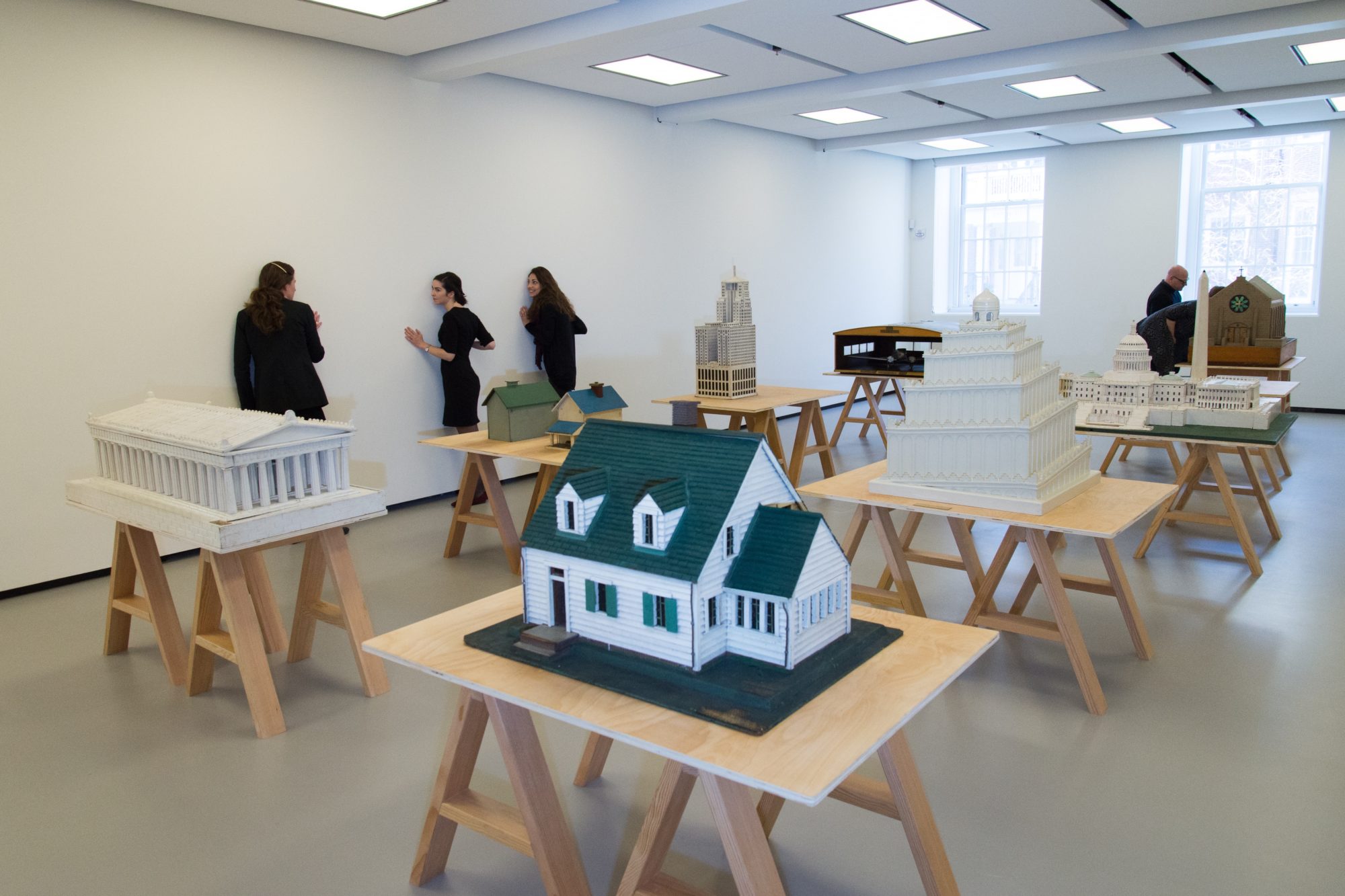 room with 8 model buildings, 3 women on a white wall and an obscured group closely viewing