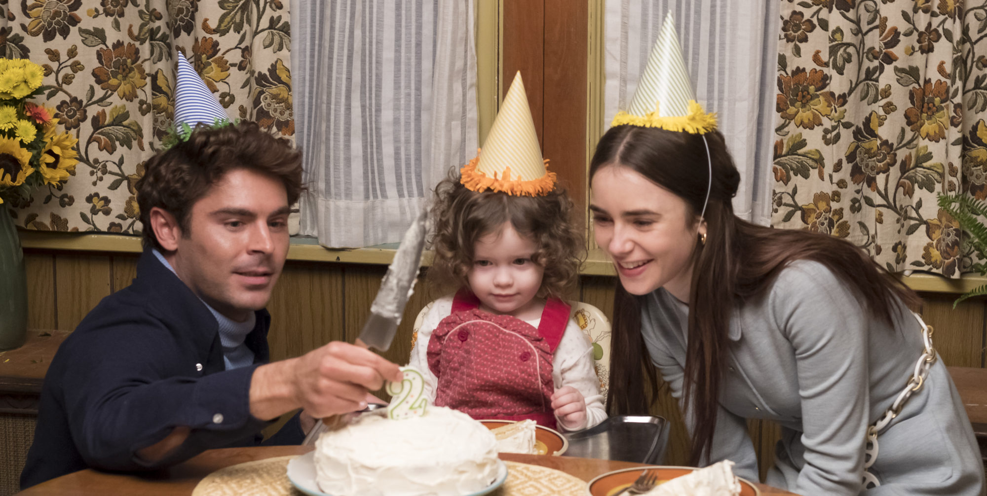 a man and a woman wearing birthday hats gathered around a birthday cake at the table with their daughter who is also wearing a birthday hat