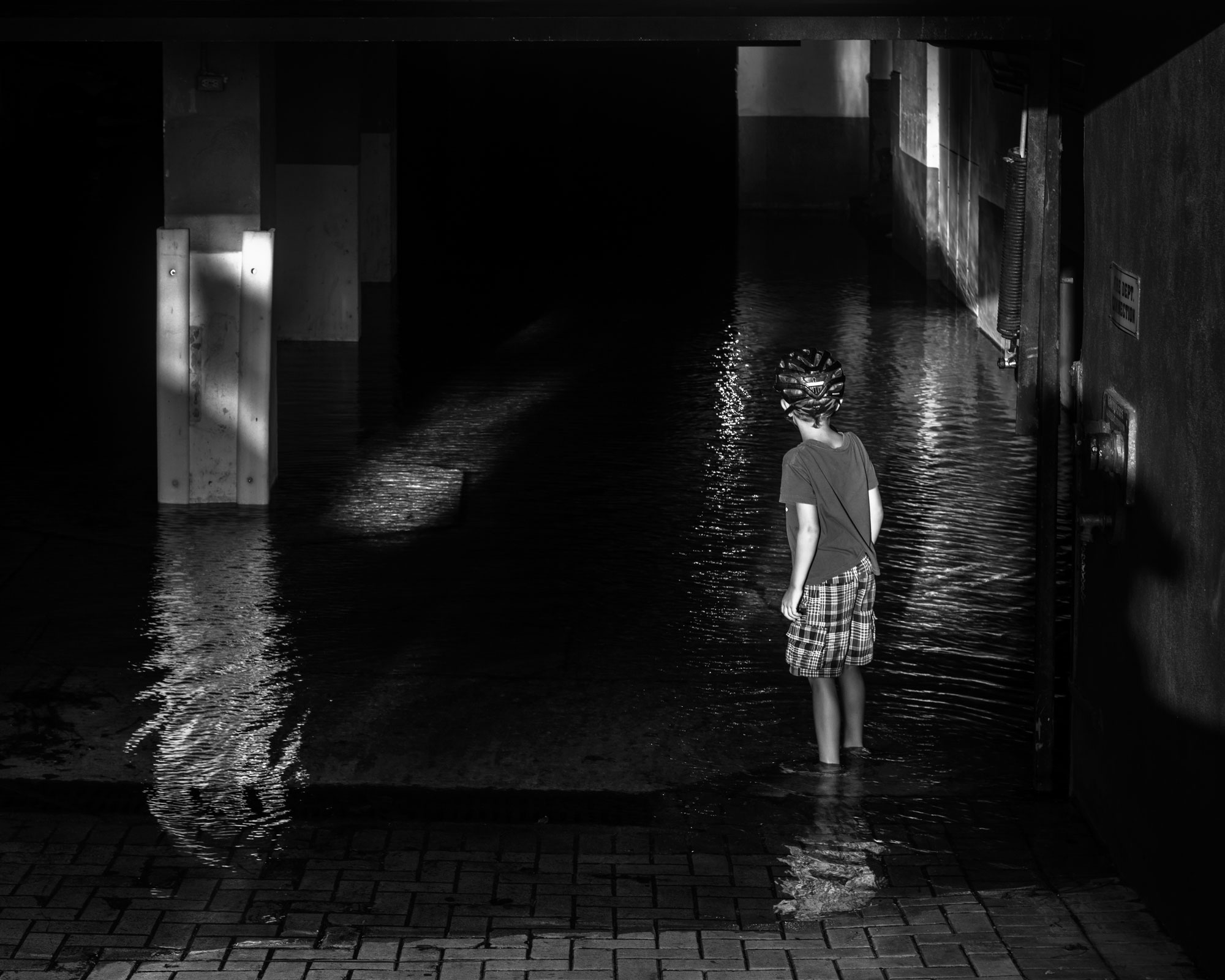 Black and white photograph of a little girl, back towards camera standing in a flooded garage. angle deep in water.