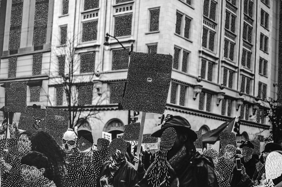 Black and white photograph of protest in city street with textured embellishments over faces and windows.