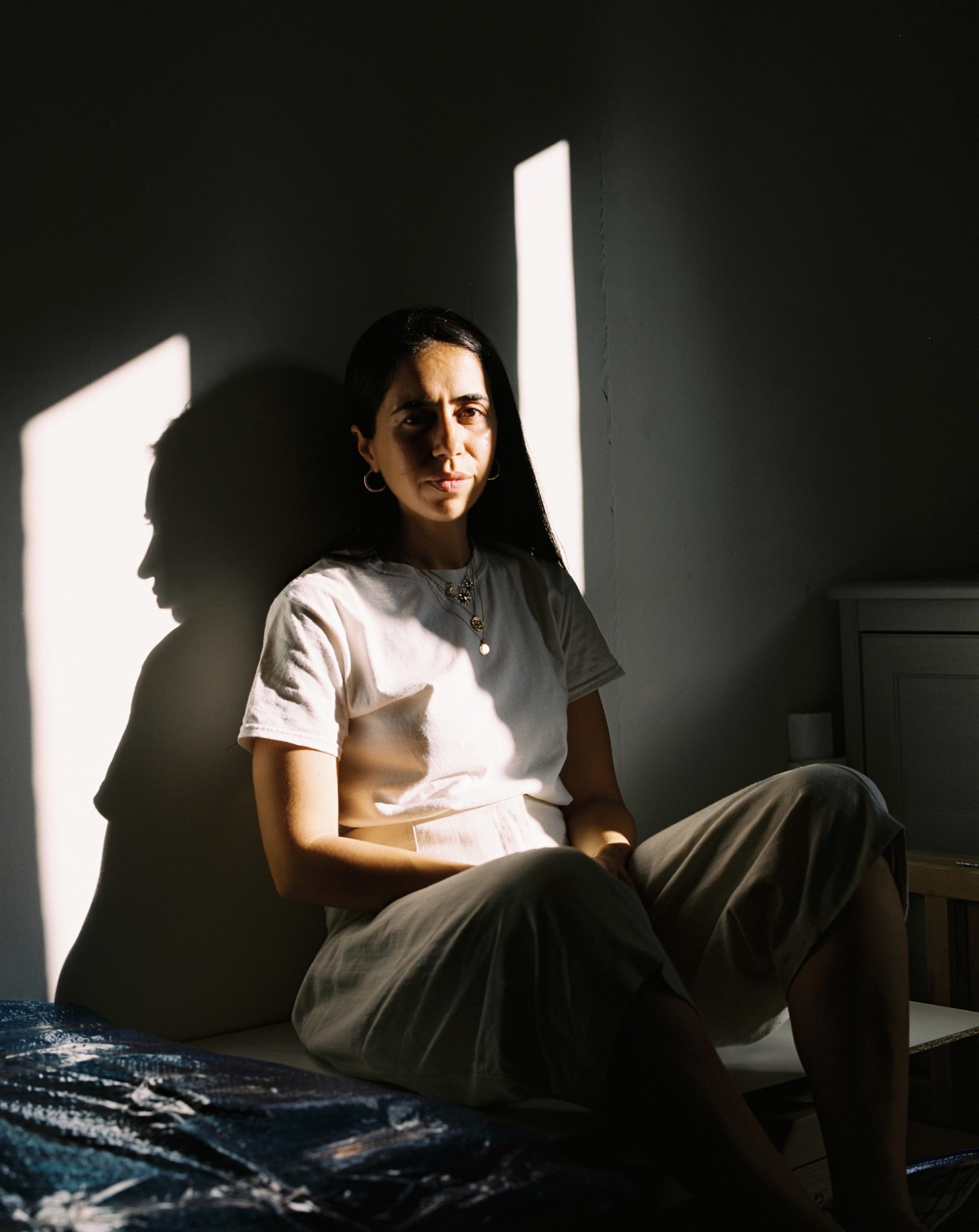 A person wearing a white shirt sits in partial light against a white wall