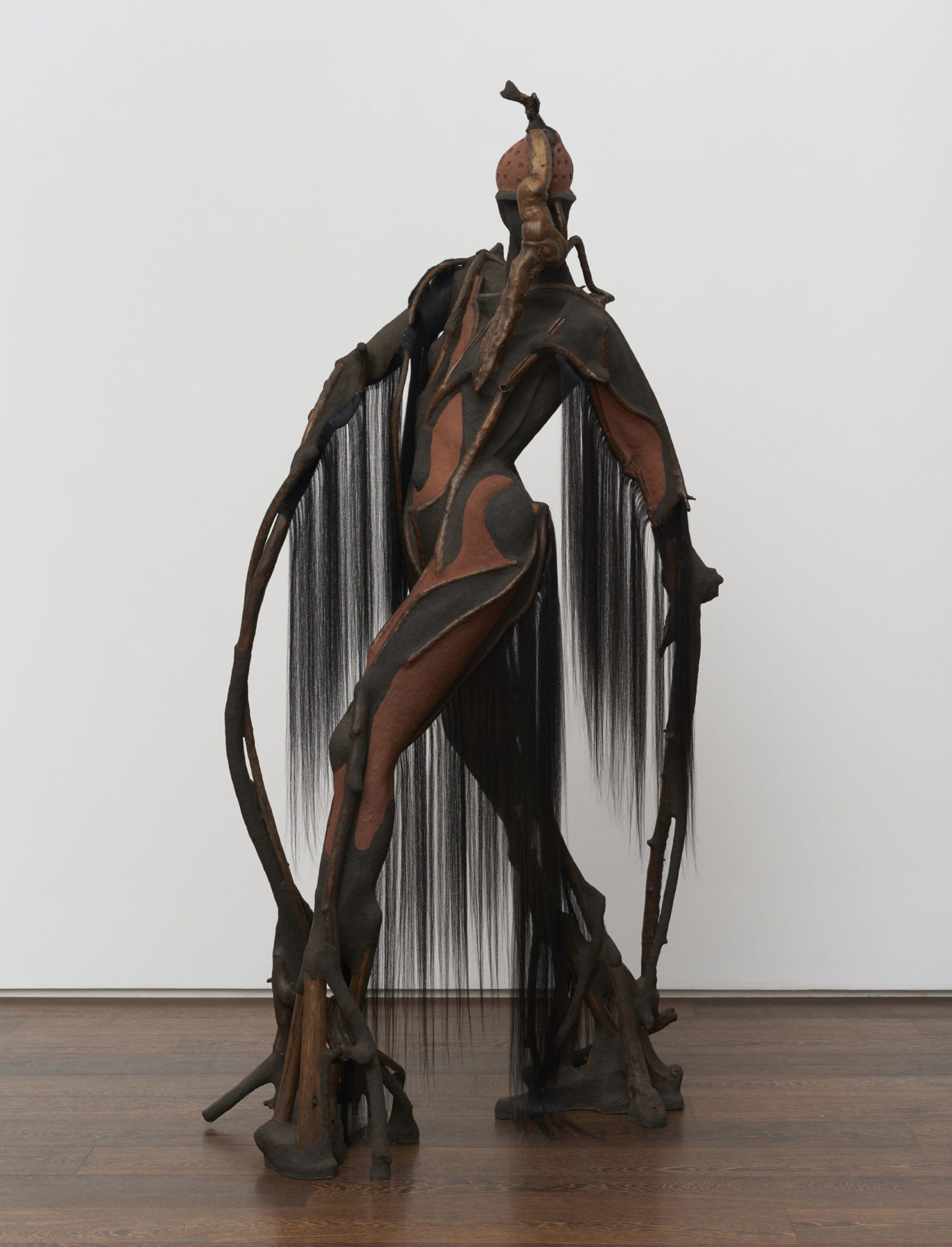 A freestanding figurative sculpture composed of dark brown wood and black fibers is set against a white wall background