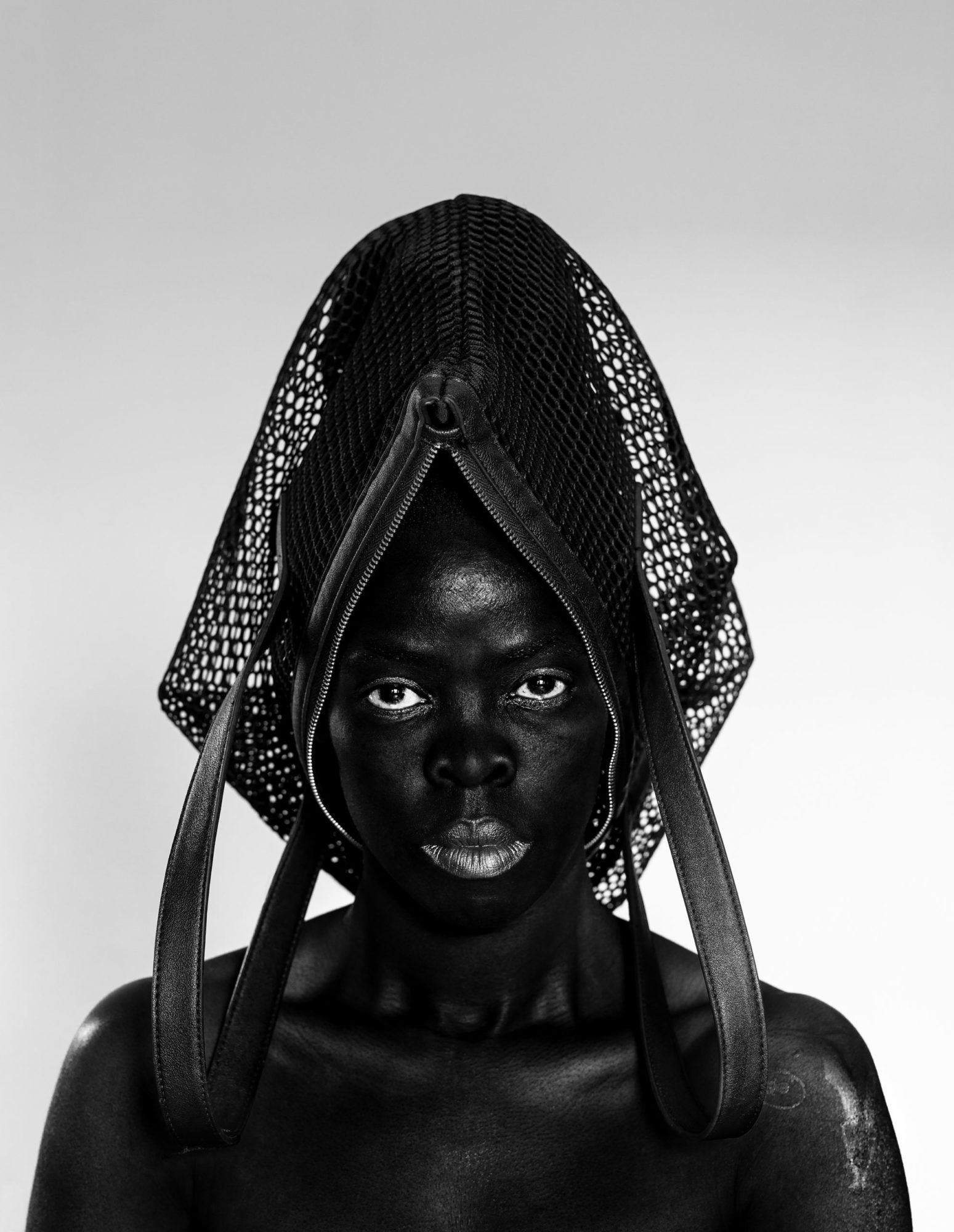 A black person with a woven headpiece stares directly out at the viewer against a very pale gray background