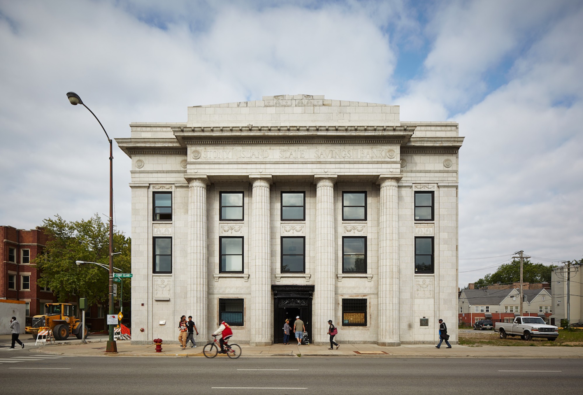 Shot across an avenue of a white greek-style bank building; the sky in the background is slightly cloudy; people are seen on the sidewalks and in the doorway