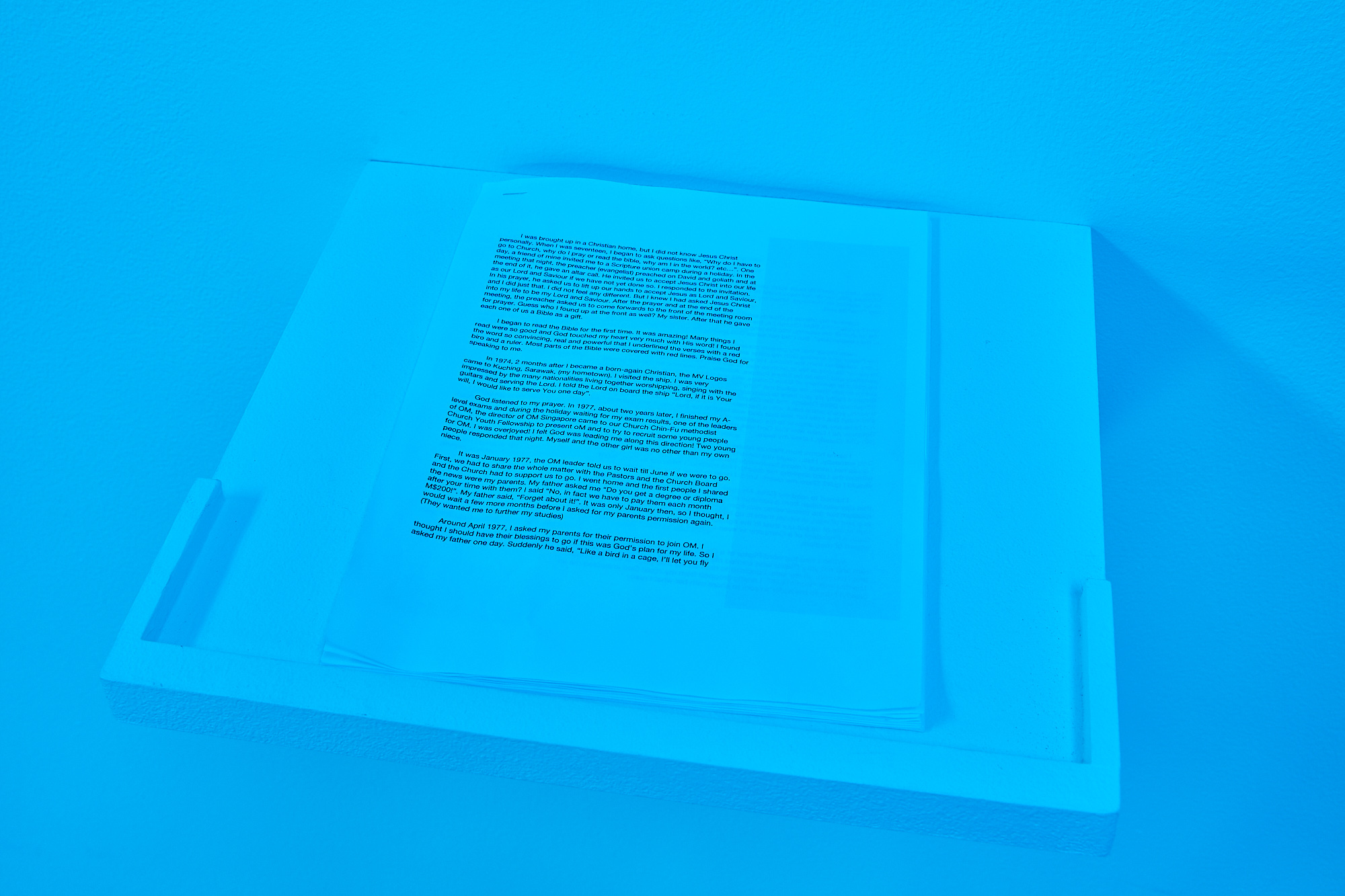a stack of computer paper with text and a staple in the upper left hand corner rests on a shelf in a blue-tinted room