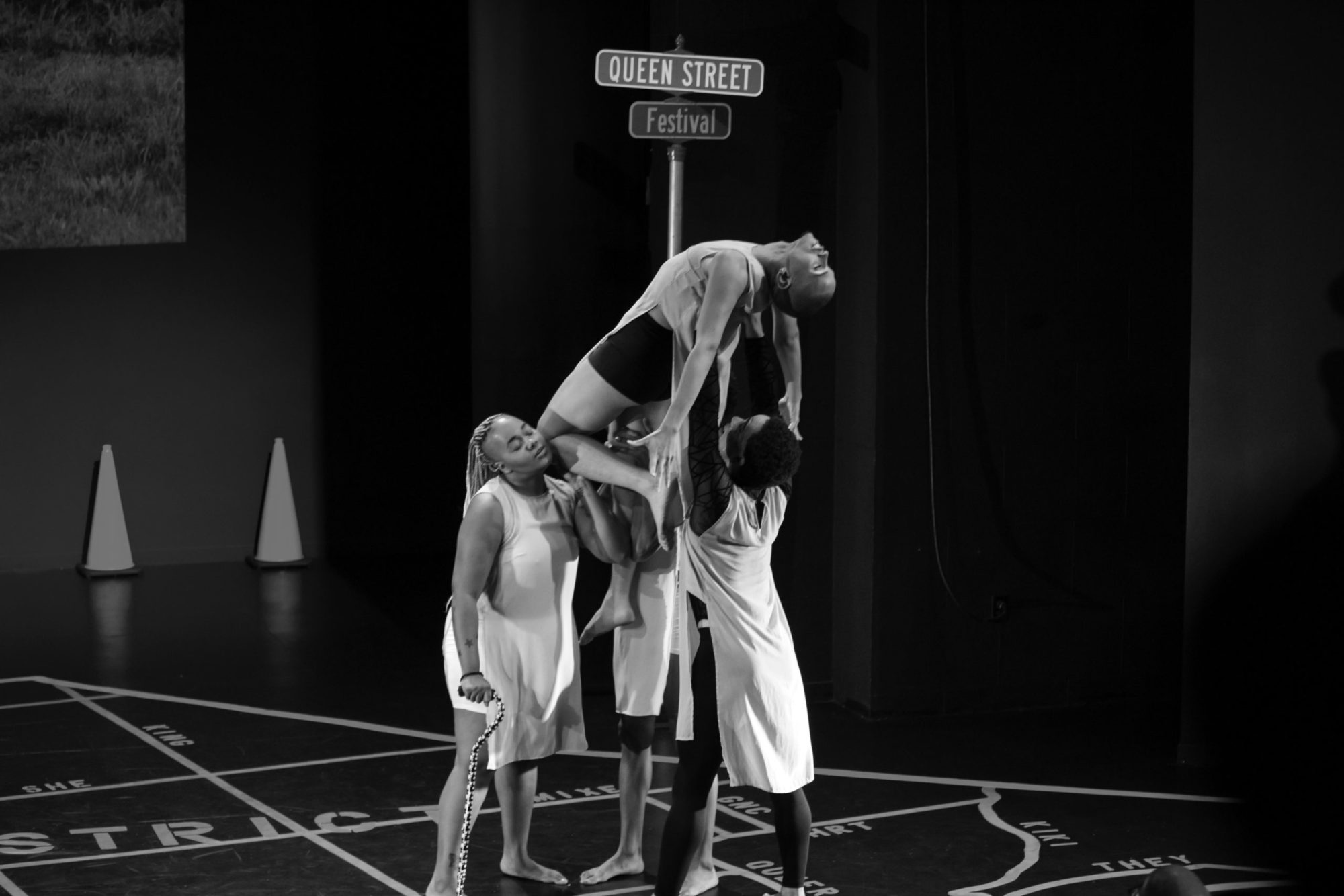 A black and white image of 3 people holding a fourth person aloft, they are standing in front of a street sign that says 