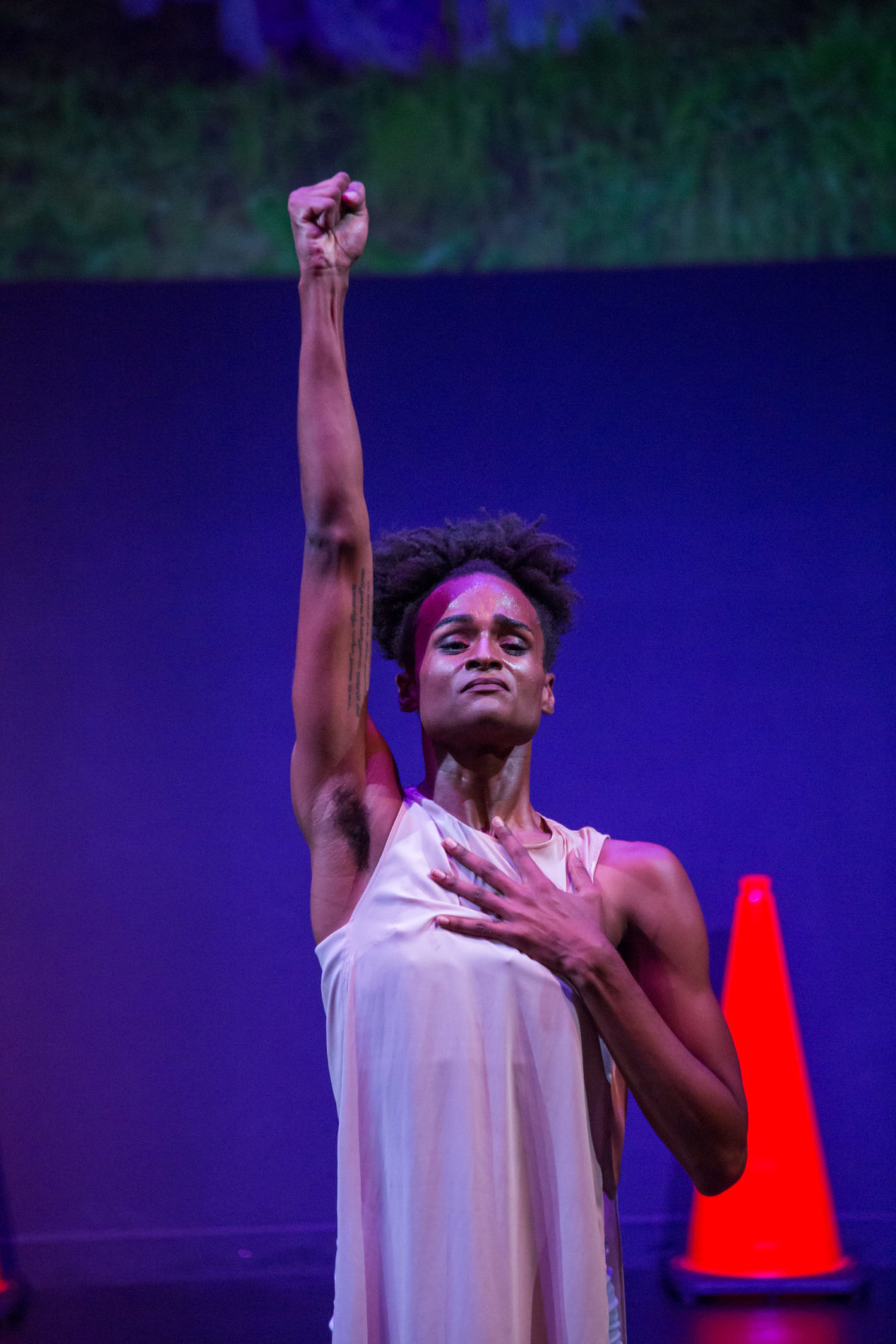 A person stands with a raised fist and open hand on their chest, they are against a blue background