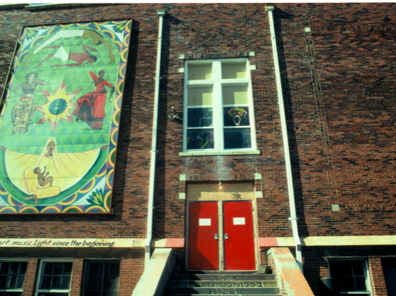 A photograph of a building exterior, with brick walls and a few stairs that lead to a bright red door. A large, colorful and mostly green poster hands on the wall, to the left of the door. Several individuals strike various poses on the poster, which has a colorful patterned border.