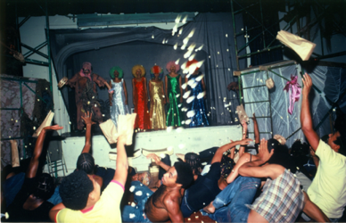 A photo of several people in a venue clumped together with their hands in the air, as if throwing something. In the back of the photograph is a stage on which five colorfully dressed individuals stand, looking out at the crowd.