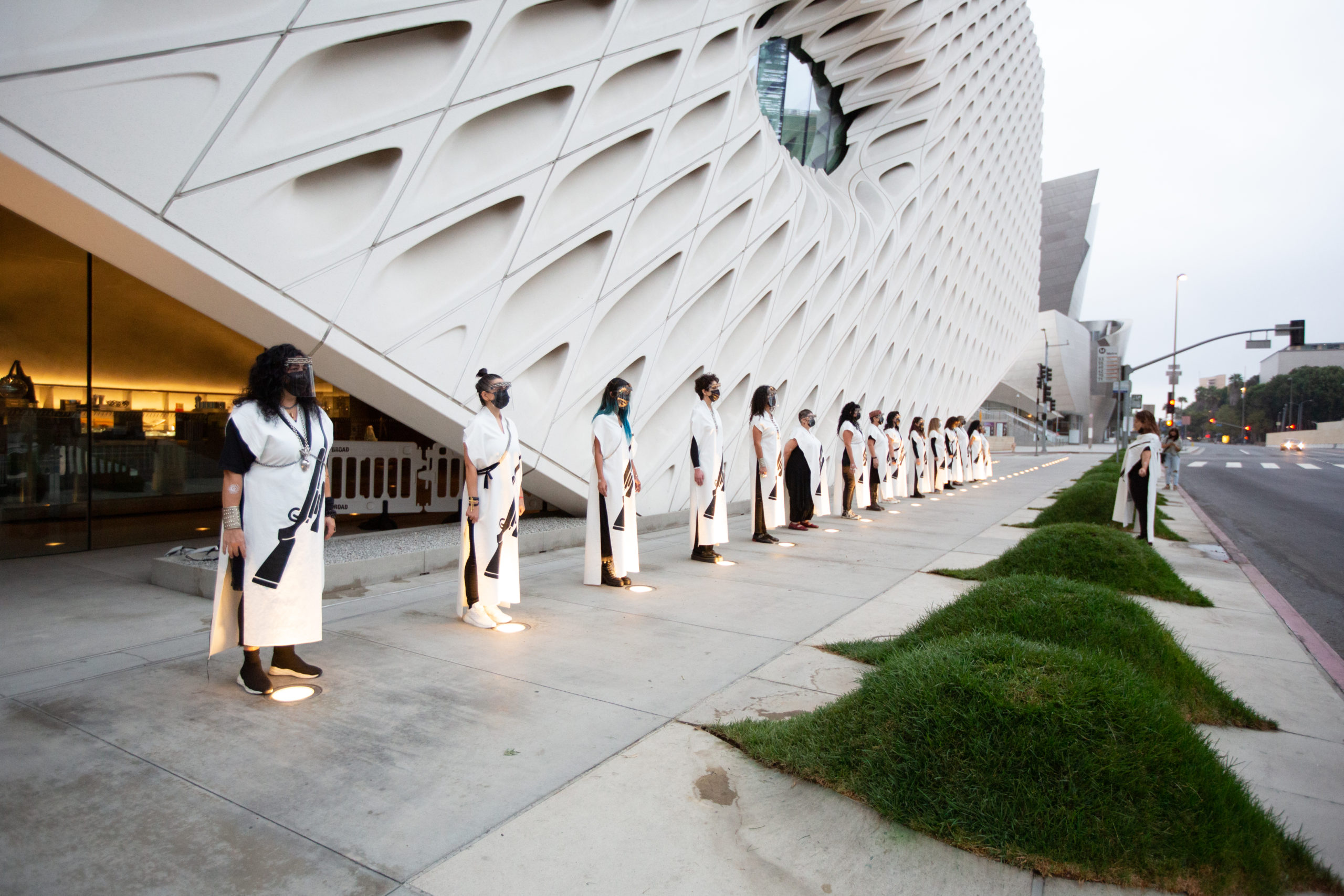 Over a dozen people lined up horizontally outside of a large building, facing one person on the opposite side of them also dressed in white and black.