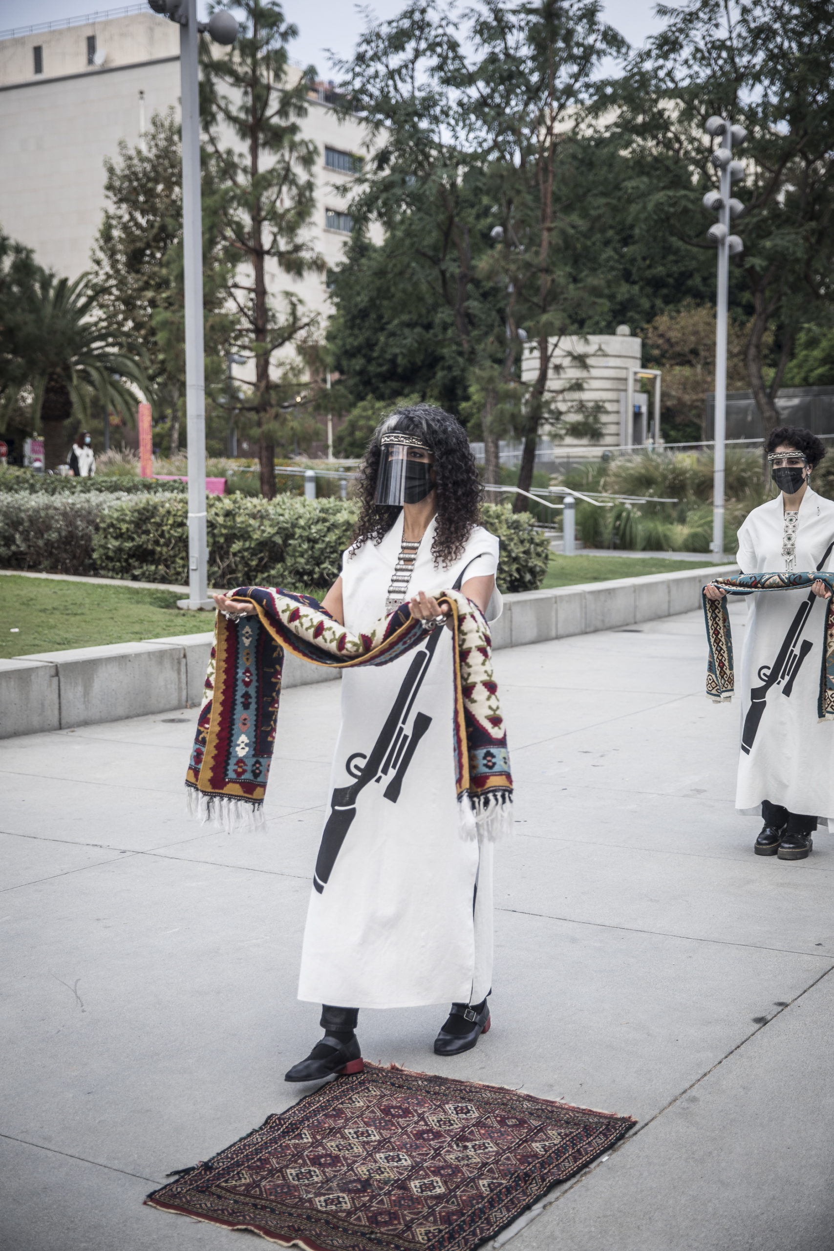Two people walking carrying small rugs in their outstretched hands, wearing white dresses with a graphic of a gun on them. A small rug is on the sidewalk.