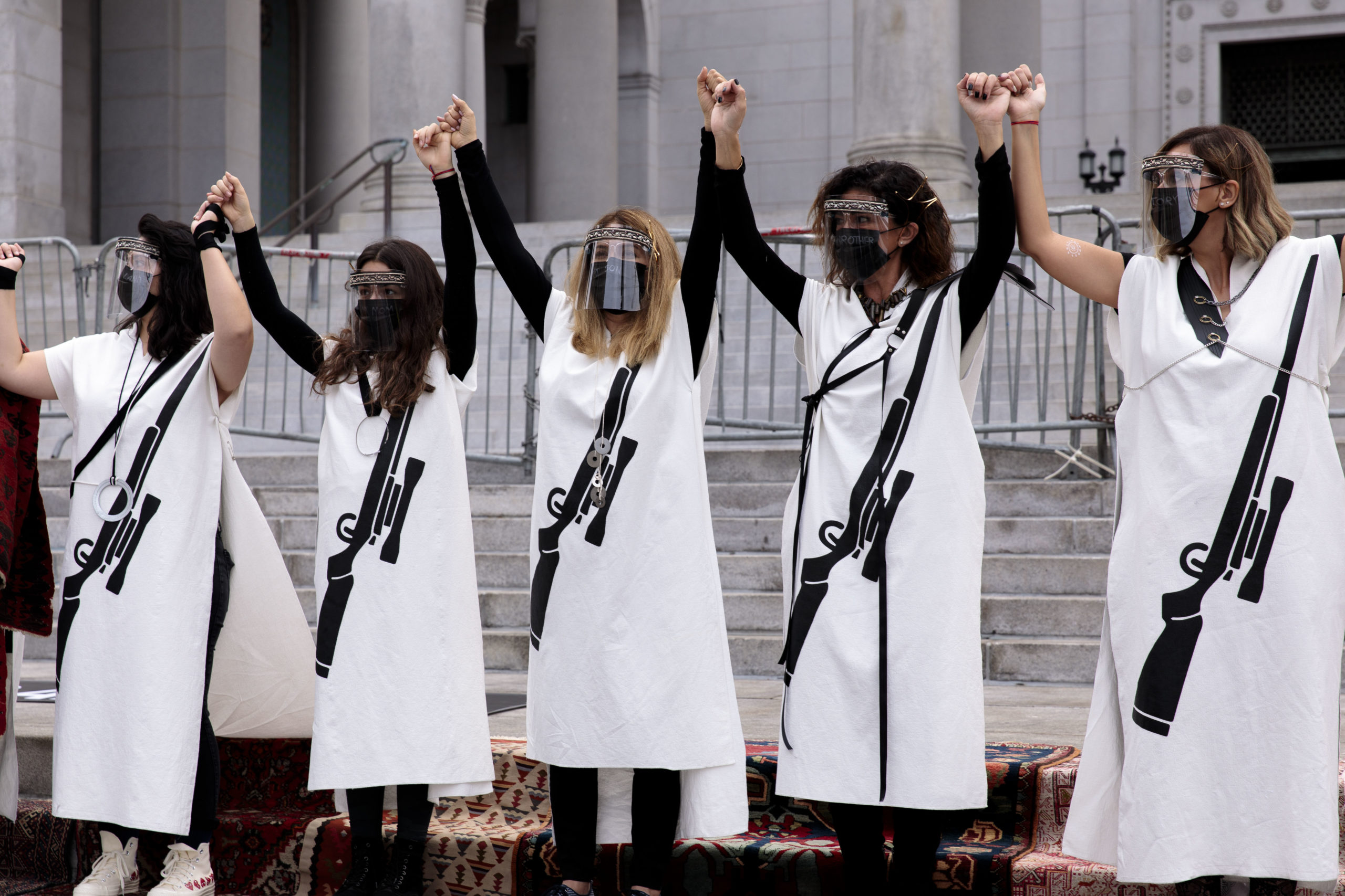 Five women wearing white dresses with a graphic of a gun on them, holding hands with their arms raised in the air.