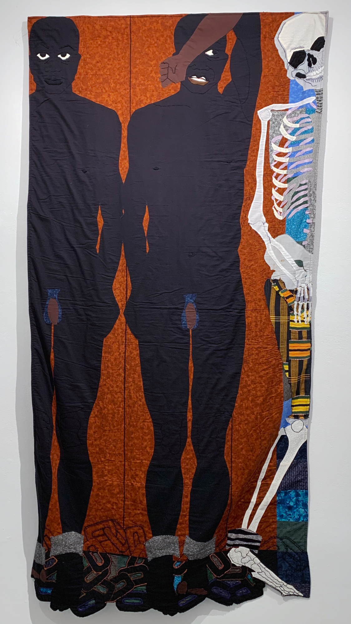 Cloth painting depicting two Black men chained together at their feet, and a human skeleton, set against a dark orange blackground
