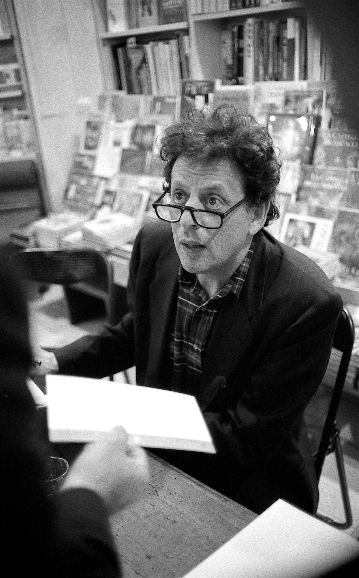 Black and white photograph of Philip Glass wearing glasses and sitting at desk in front of bookshelf, a hand with sheet of paper reaches into frame