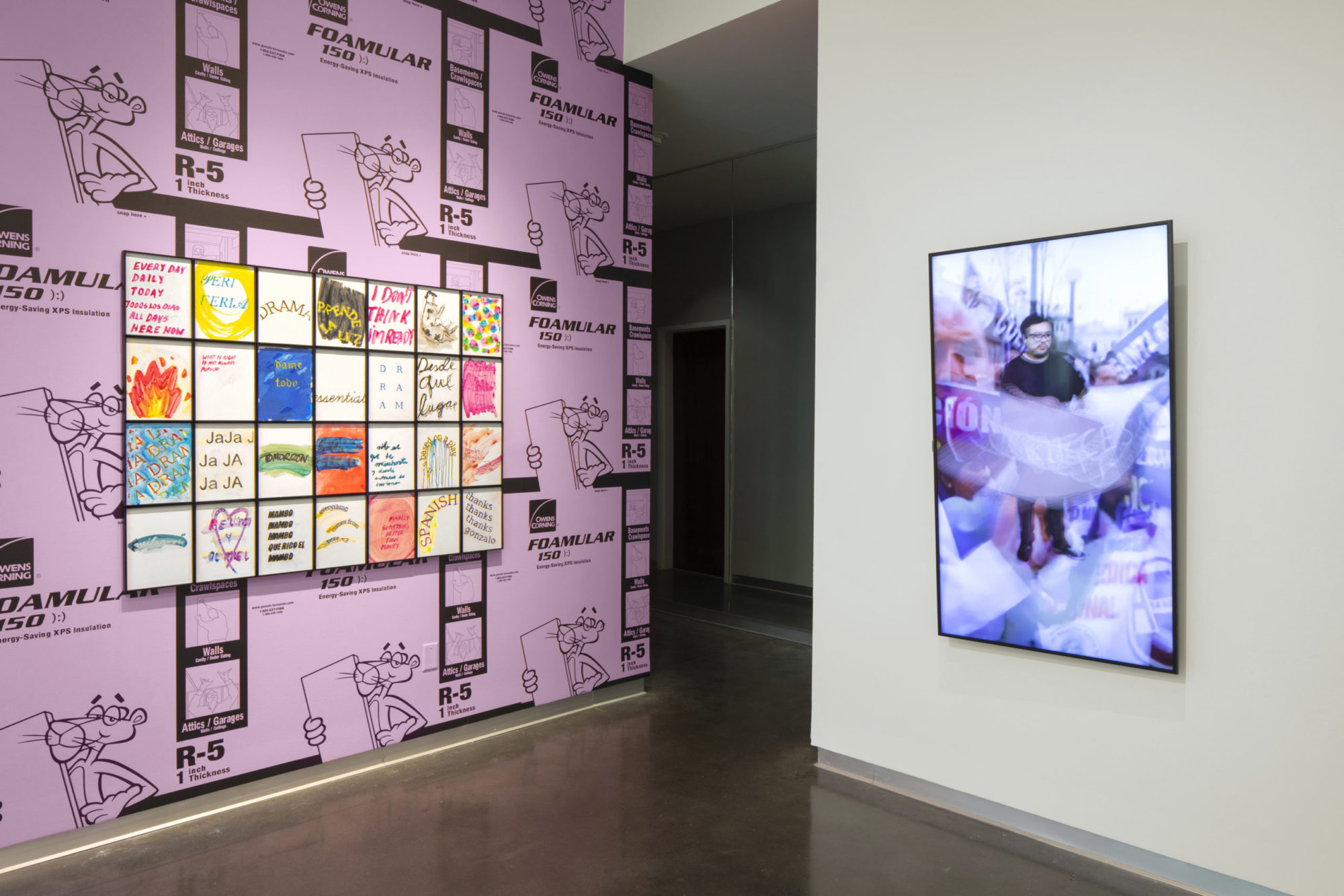 Wall covered in Pink Panther insulation foam hung with grid of small mixed-media works printed and painted with brightly colored abstract designs and text in Spanish and English, white wall with photograph of man standing on street surrounded by blur of hand-held signs, streetlamps, and American flag