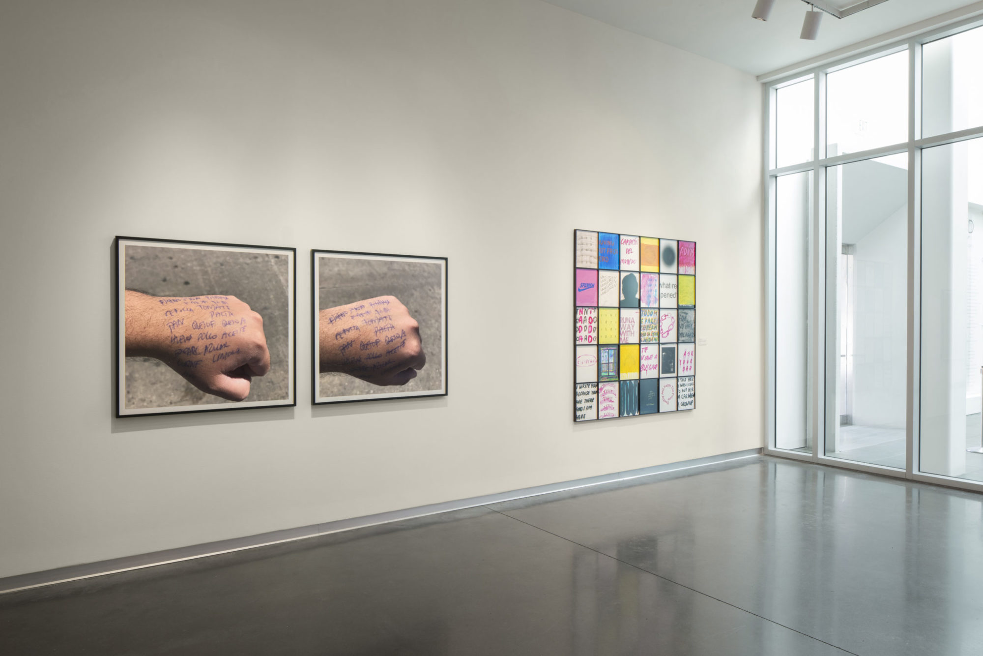 Corner of gallery with three wall-hangings, two large photographs of grocery list written on the back of a hand, grid of small mixed-media works printed and painted with text and abstract designs in black, bright pink, orange, and green