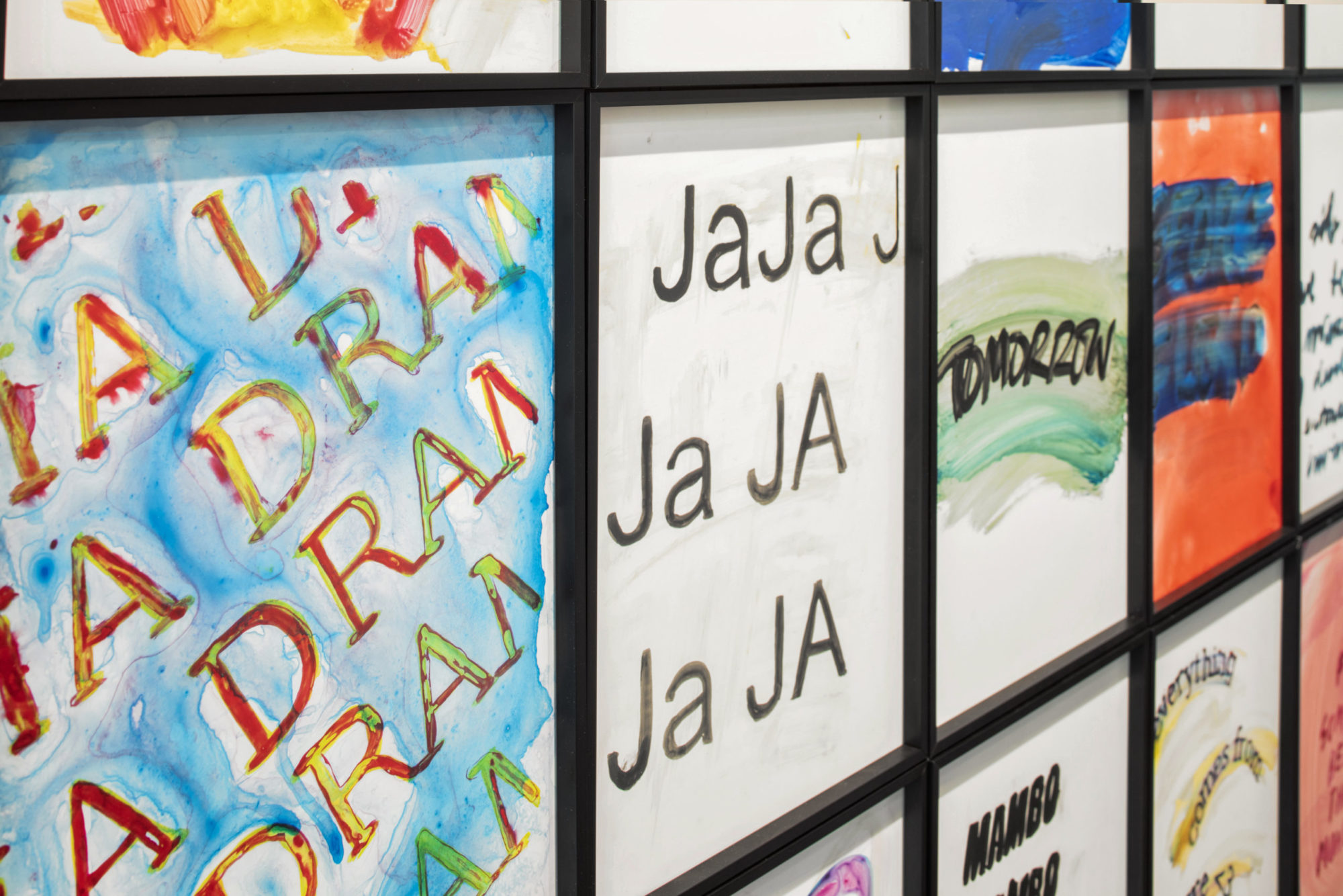 Close-up image of grid of mixed-media works combining brightly-colored, abstract, painted designs, and text in Spanish and English, including the words 