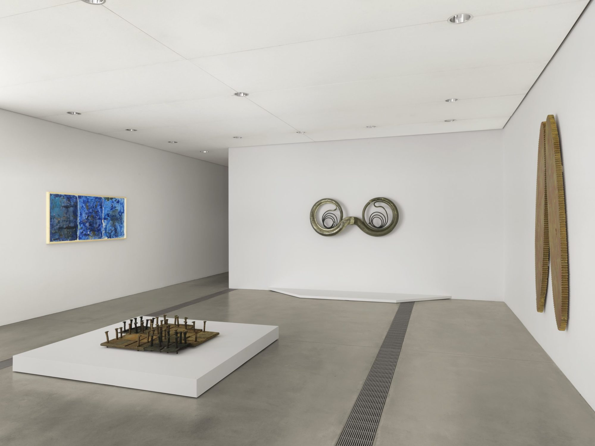 There are four visible artworks displaced. On the front facing wall is a pair of metallic bells attached to each other forming an ouroboros. On the left wall is a blue three-paneled abstract paintings that are framed in light wood. On the low center podium is a collection of steel nails that are back and brown facing downward. The nails are standing on patches of black and brown steel. On the right wall hangs a mvet (pair of connected oval shaped pieces of wood).