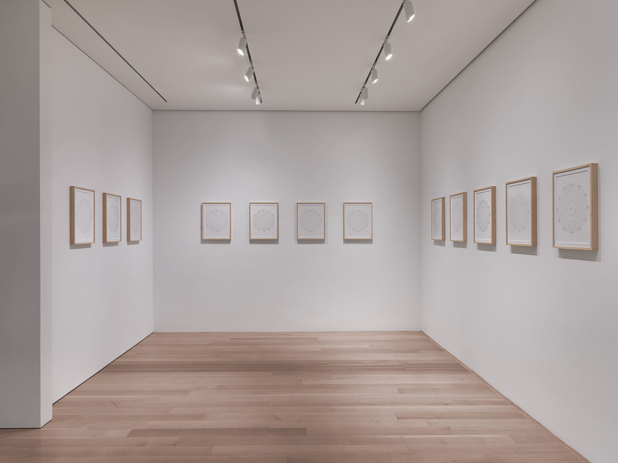 An open atrium lit by two rows of overhead lights in a gallery, with three separate walls each with framed drawings of depicting circles. The center front-facing wall has four separate framed images. On the left wall there are three images in the same fashion, and on the right wall there are five. All equally spaced out on al three walls.