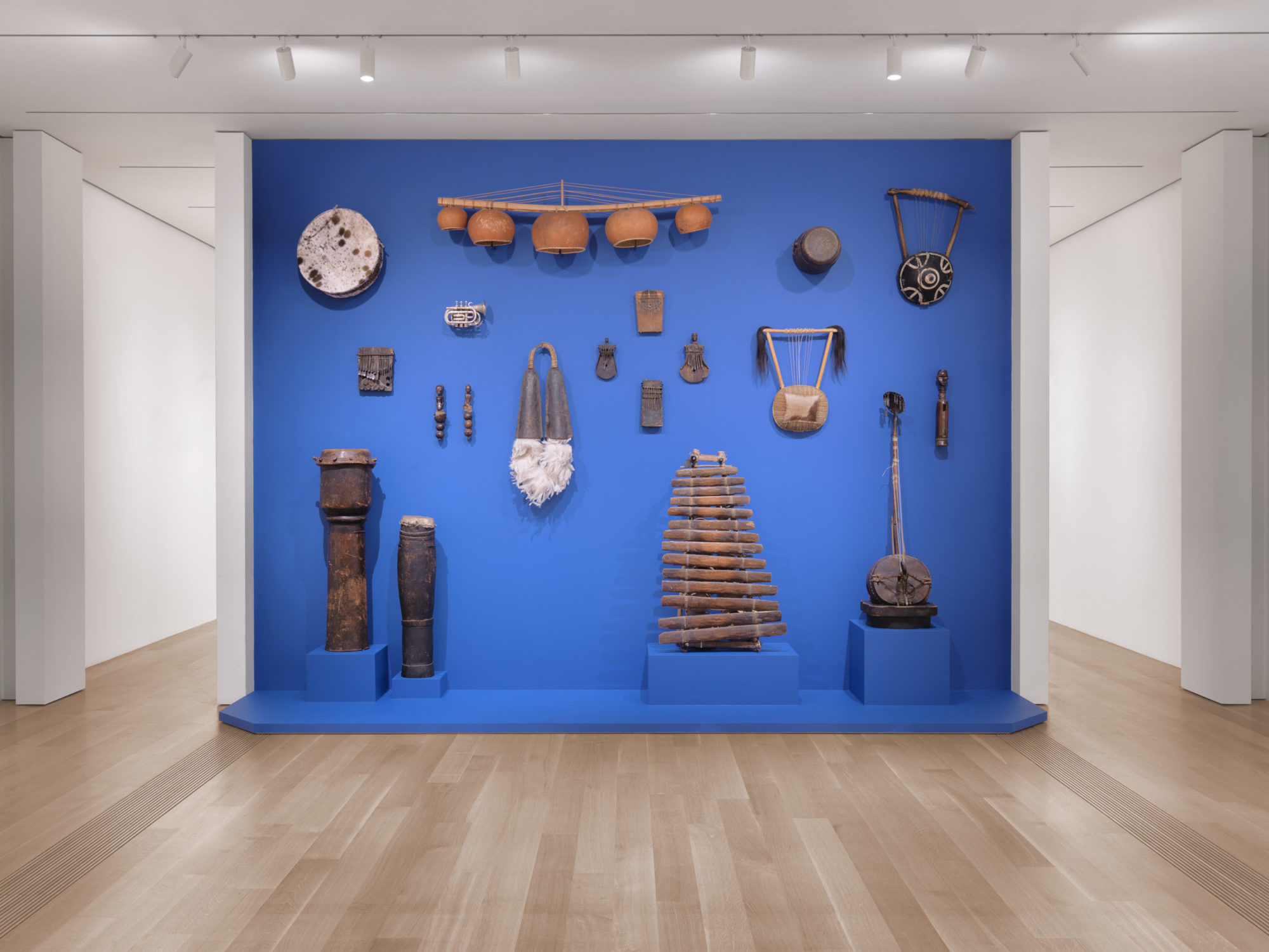 A light blue stage and backwall standing between two white columns. There are various artworks hanging on the wall and standing on podiums. The four sculptures standing each have their own podium and all are instruments made of wood, reptile skin, and hide. Two on the right side are tall cylinder drums. On the left side there is a collection of wooden logs standing upright like a xylophone. To the left of this is a banjo with a long neck. Hanging on the wall are several instruments and accompanying tools including thumb pianos, bowl lyre, and harp lute.