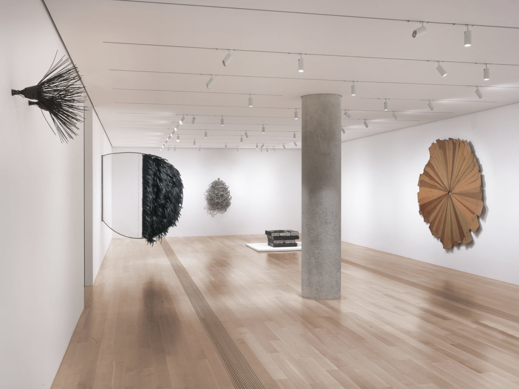 Open gallery room with large gray stone column in the center. Artwork on each wall, one piece hanging above ground on each wall. To the right, we see two pieces, one closer to the camera which is two black straw-like brissles facing outward from the wall and the other piece further down the same wall is a large black feathered circular structure that is protruding from the wall by a wire. The black feathered body is not touching the wall. On the front facing wall there is a distant gray sculpture hanging up. It is a cluster of aluminum clothing hangers. To the left wall, there is a large flattened circle of wooden planks. It is a variety of different shades of brown, and various lengths.