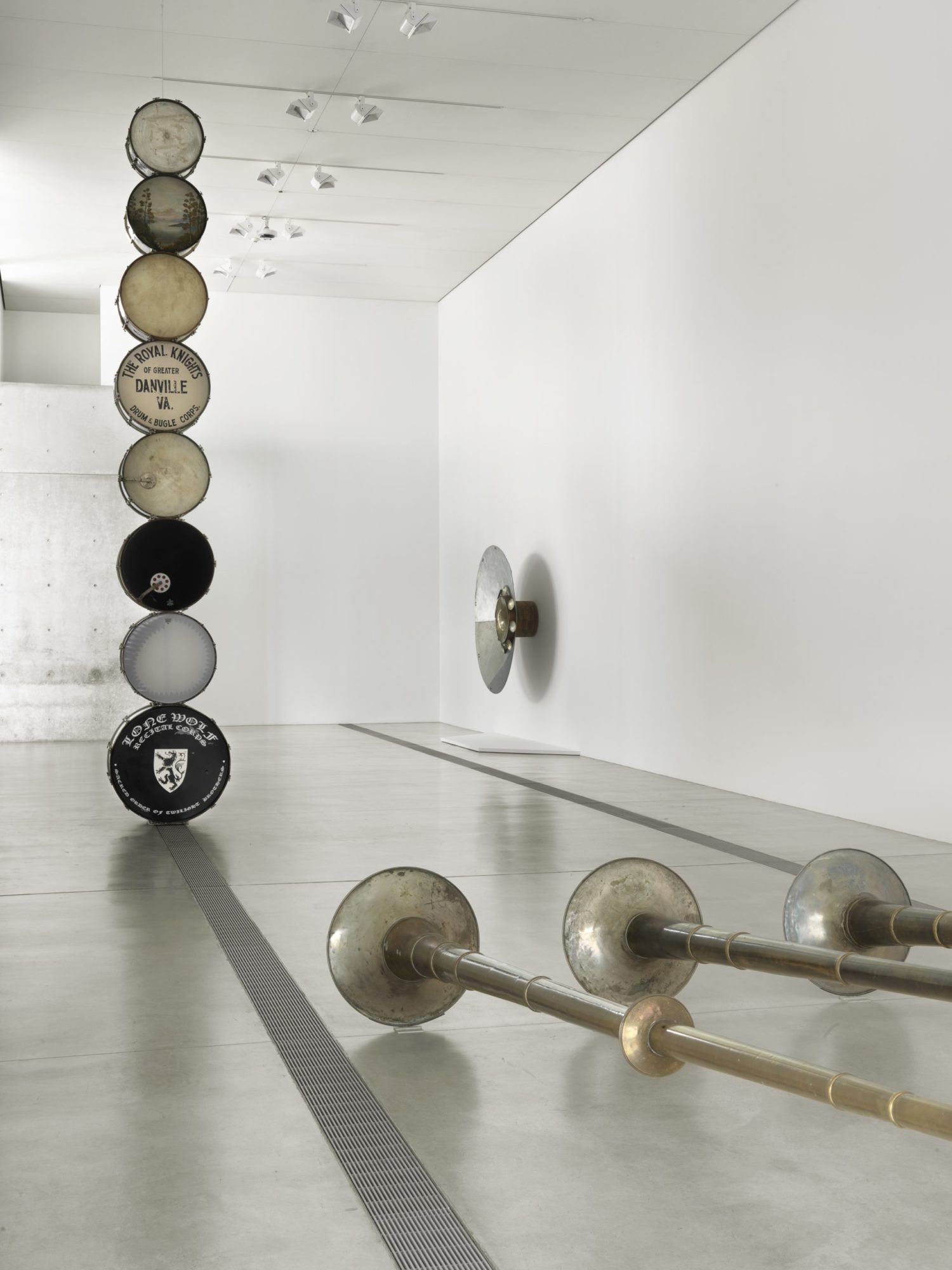 A large column of eight drums in the center of a white room. They are standing on their side, with the drumhead facing the camera. There is a variety of colors such as black drums, white, and light brown.