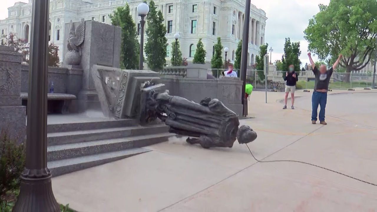 Toppled over statue of Christopher Columbus on the ground, with three people nearby looking and cheering.