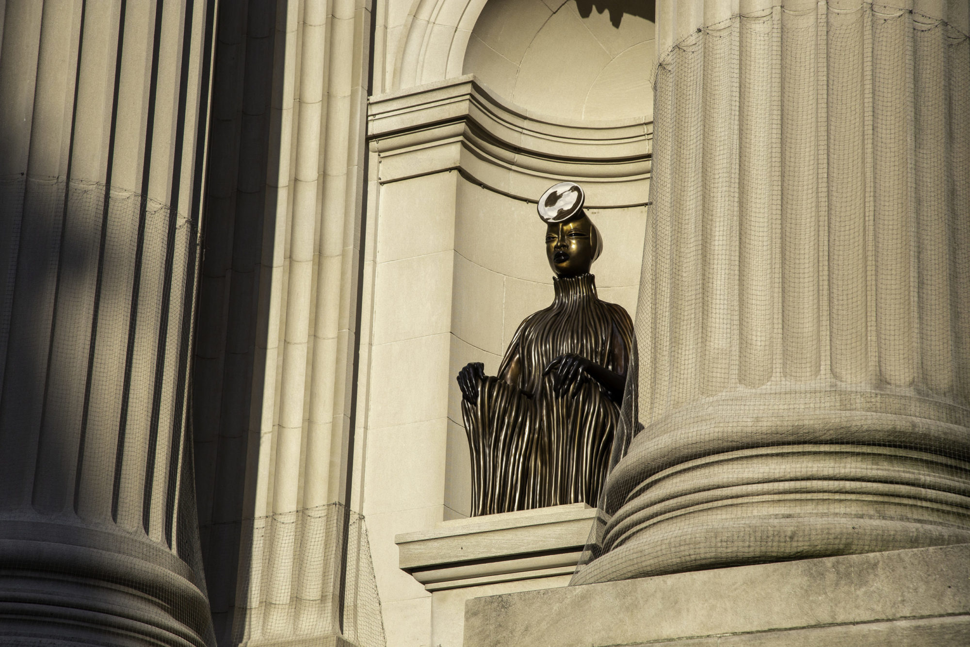 A wide image of the sculpture's left profile, lit up by sunlight in between two columns. The face is visible on the seated sculpture, as well as their circular headpiece.