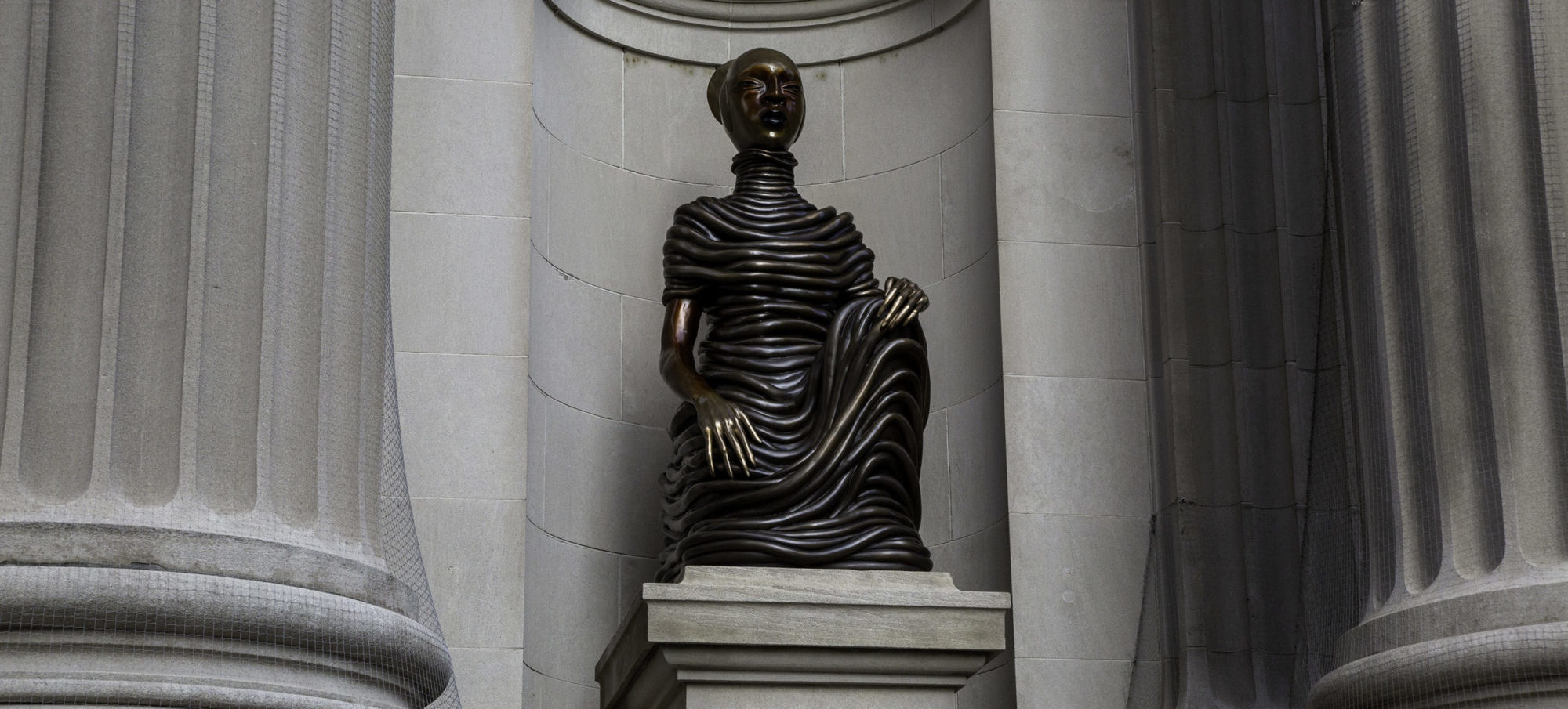 Front facing image of a sculpture: a figure kneeling with one arm in lap and another raised. The figure has rings wrapped around it. It sits in between large columns and looks straight ahead.