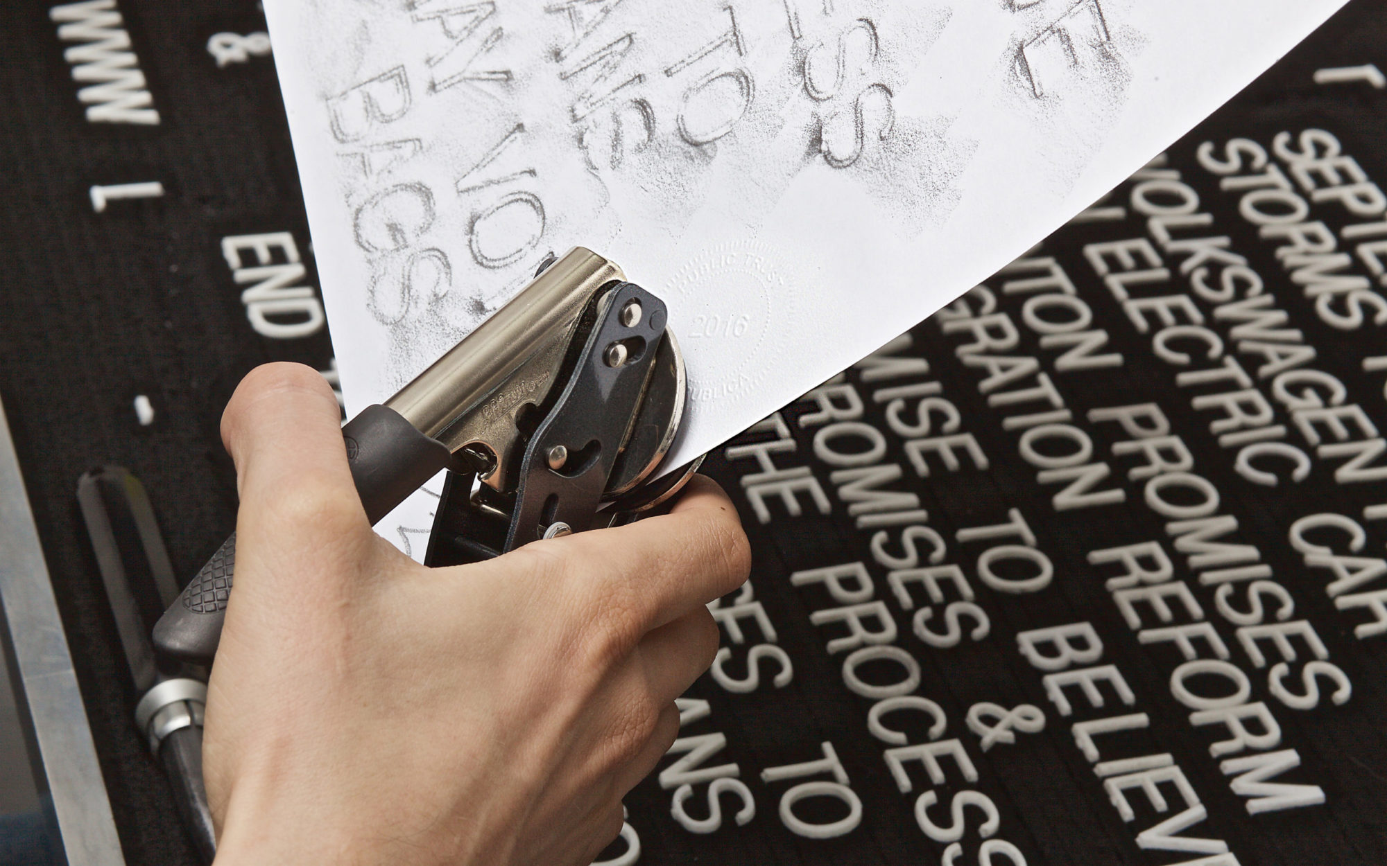 Close-up of hand stamping paper with metal embossing tool, black letterboard with white letters spelling words including 