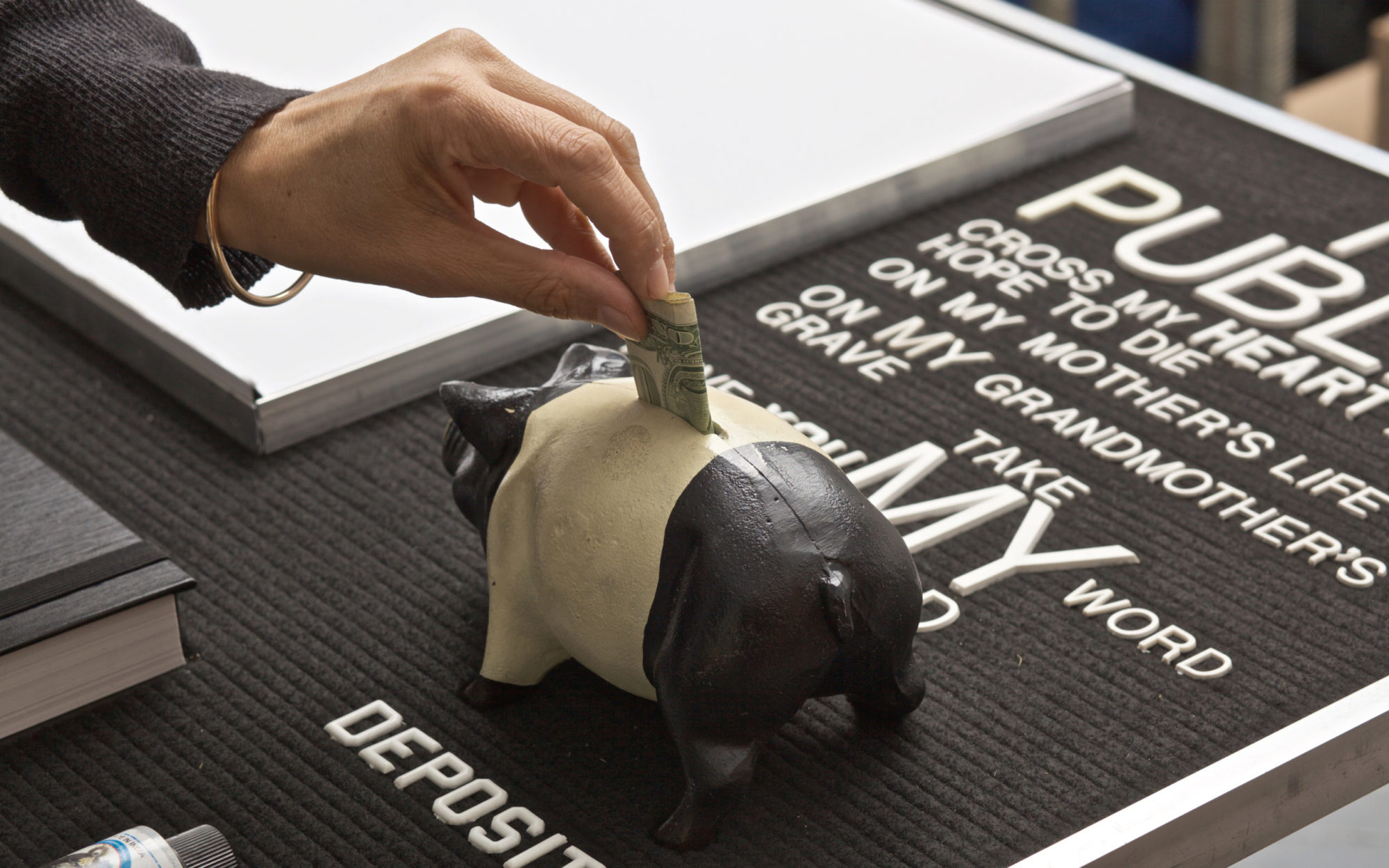 Hand stuffs folded U.S. currency into black and white plastic pig, pig stands on black letterboard with white letters spelling various commonly used phrases signifying a promise